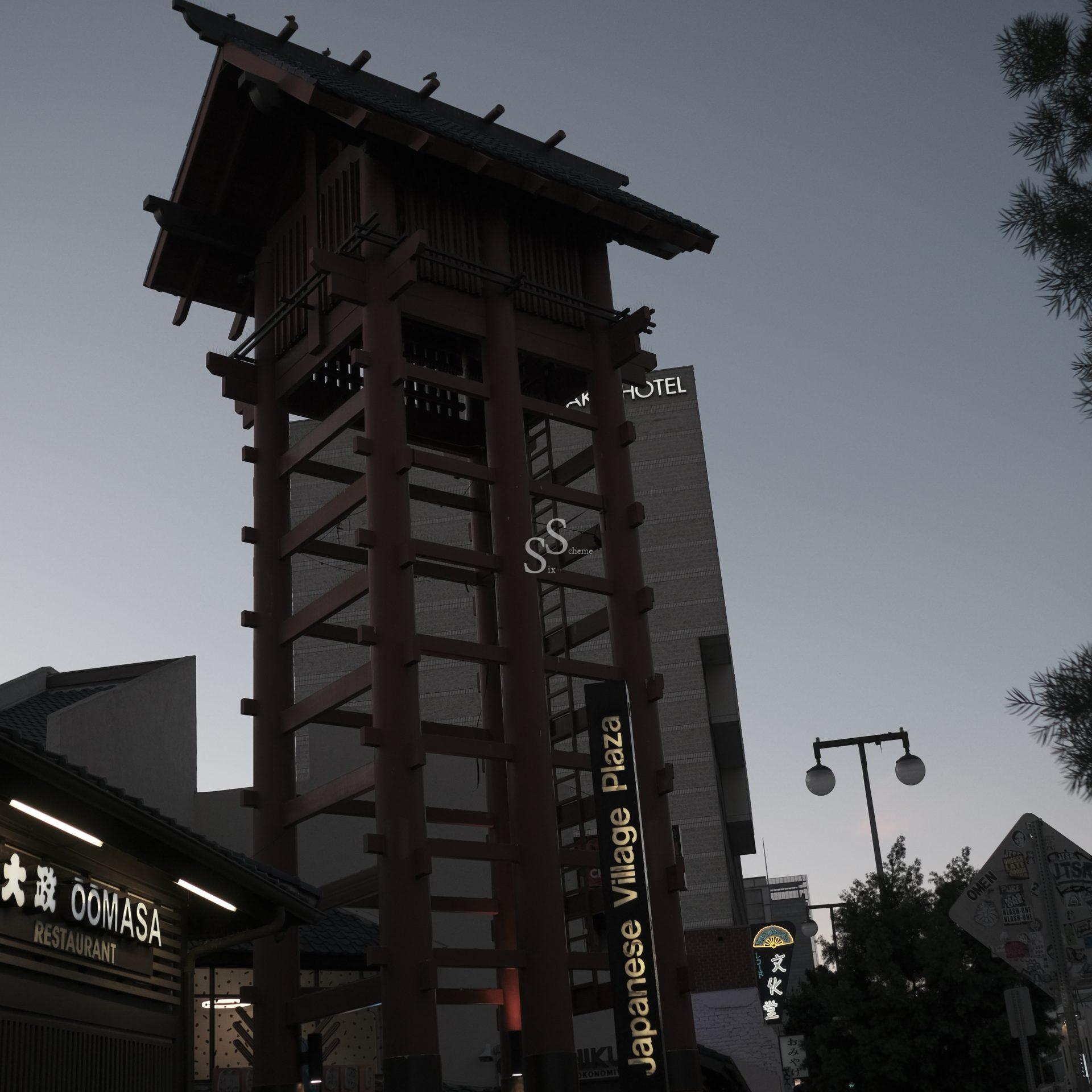 A tall wooden tower stands in Japanese Village Plaza at dusk, with restaurant signs in English and Japanese below and nearby trees and modern buildings in the background.