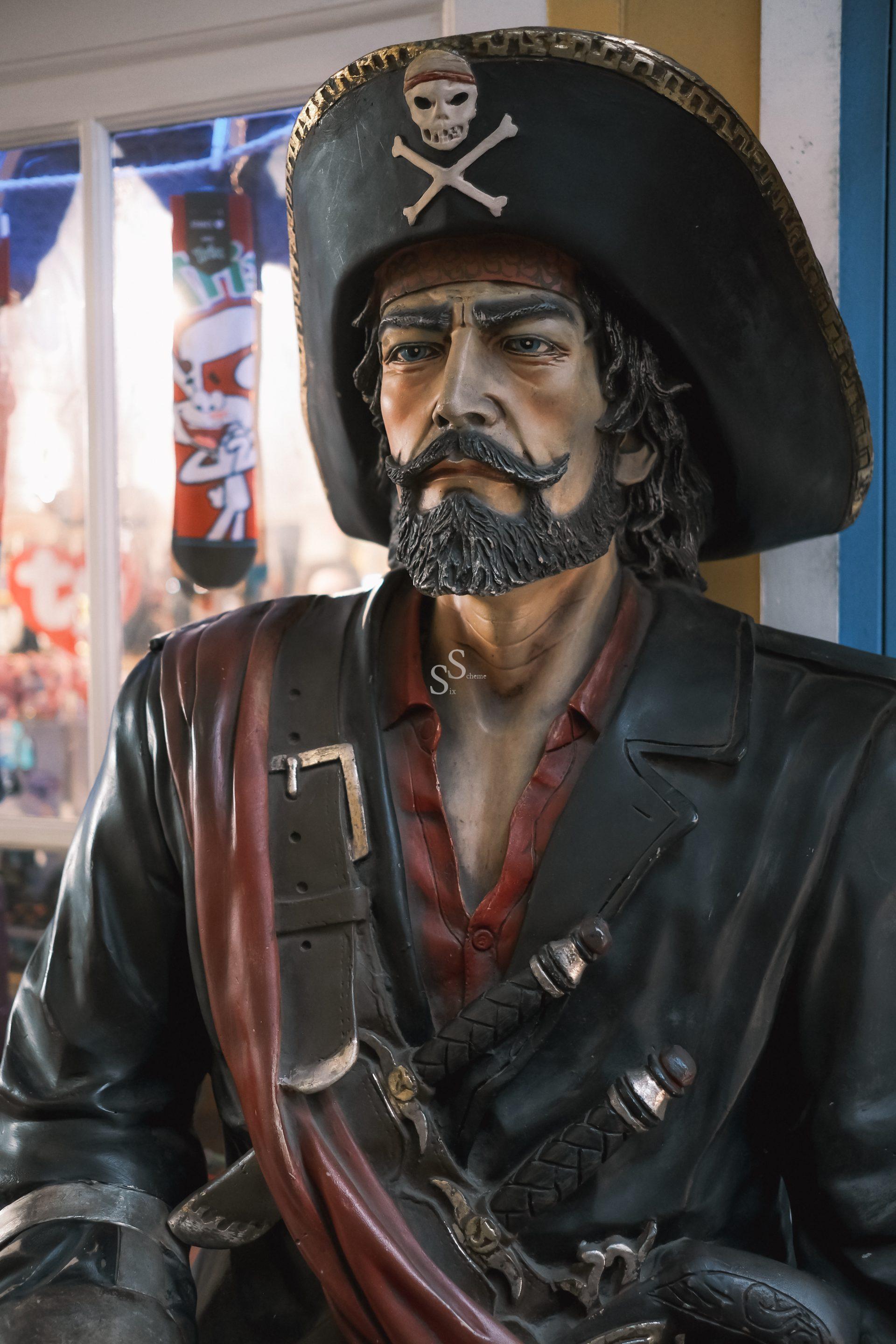 A statue of a stern-looking pirate with a black hat featuring a skull and crossbones, a red sash, and swords across his chest, standing indoors near a window.