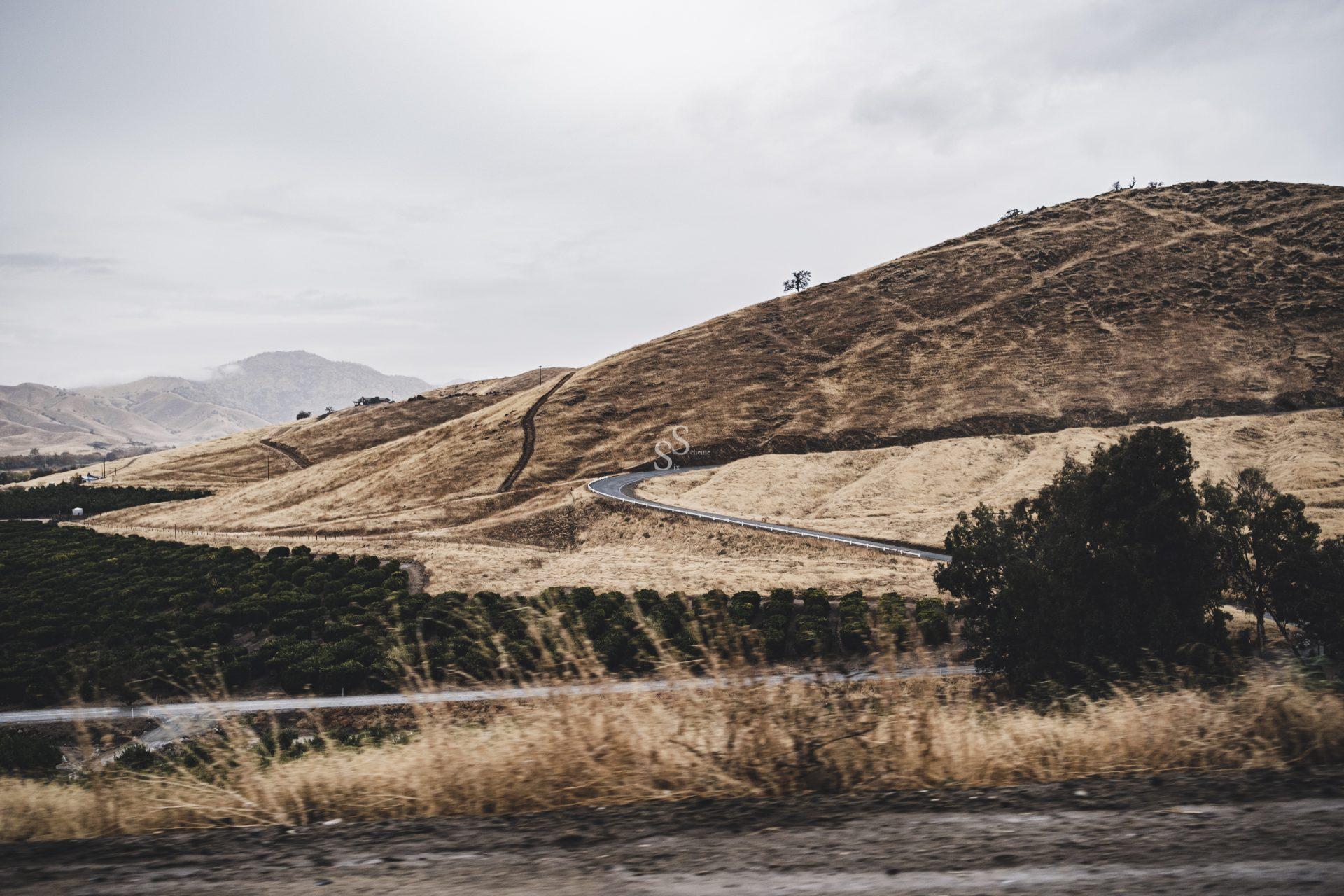 A winding road curves through dry, grassy hills under a cloudy sky, with green trees and fields in the foreground.