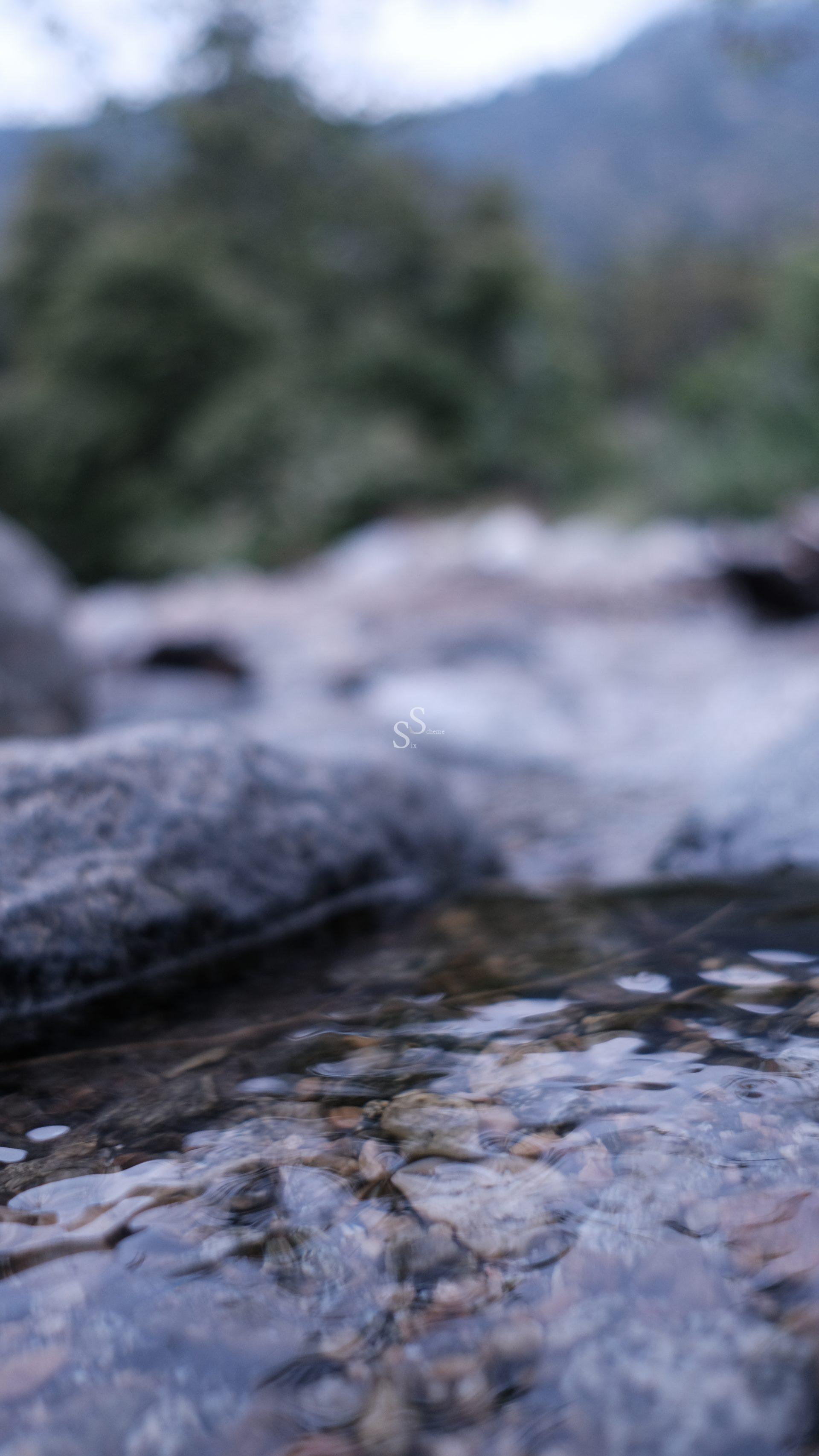 A close-up view of a shallow stream flowing over rocks, with the background blurred and out of focus, suggesting a natural outdoor setting.