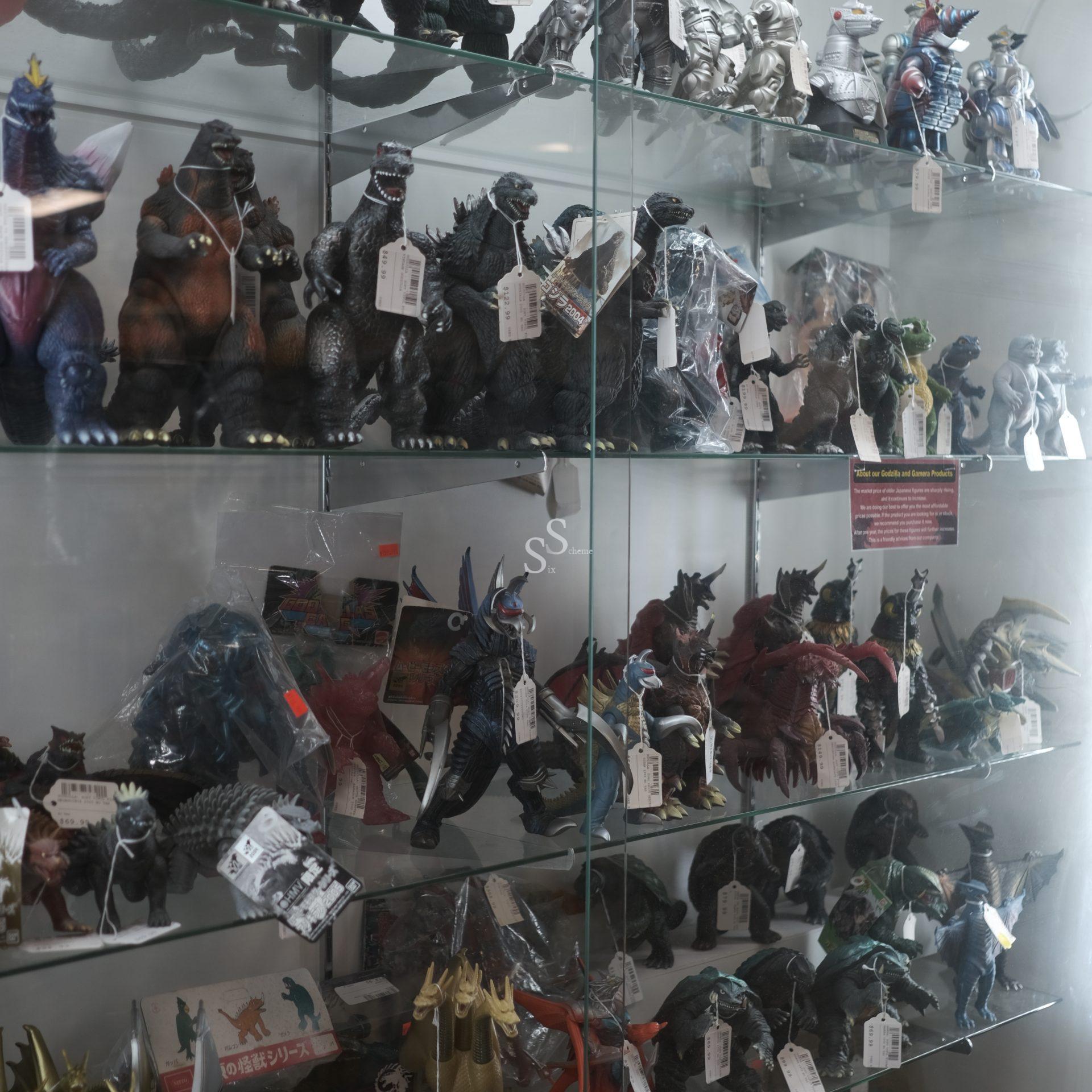 A glass display case filled with various Godzilla and kaiju action figures, each with price tags, arranged on multiple shelves. The figures vary in size, color, and pose.