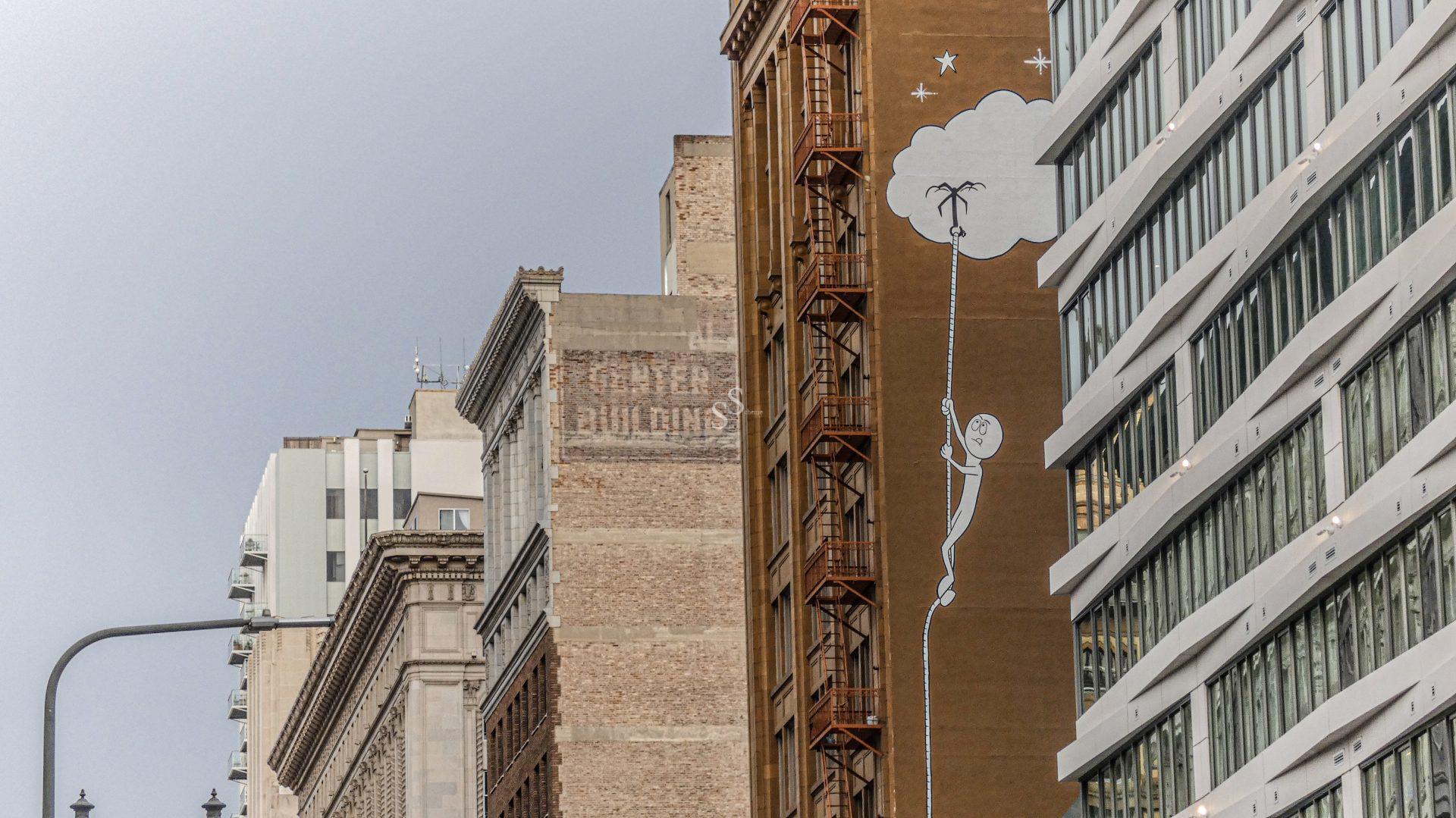 A city street with tall buildings; one building features a mural of a stick figure climbing a rope to a cloud to reach the stars.
