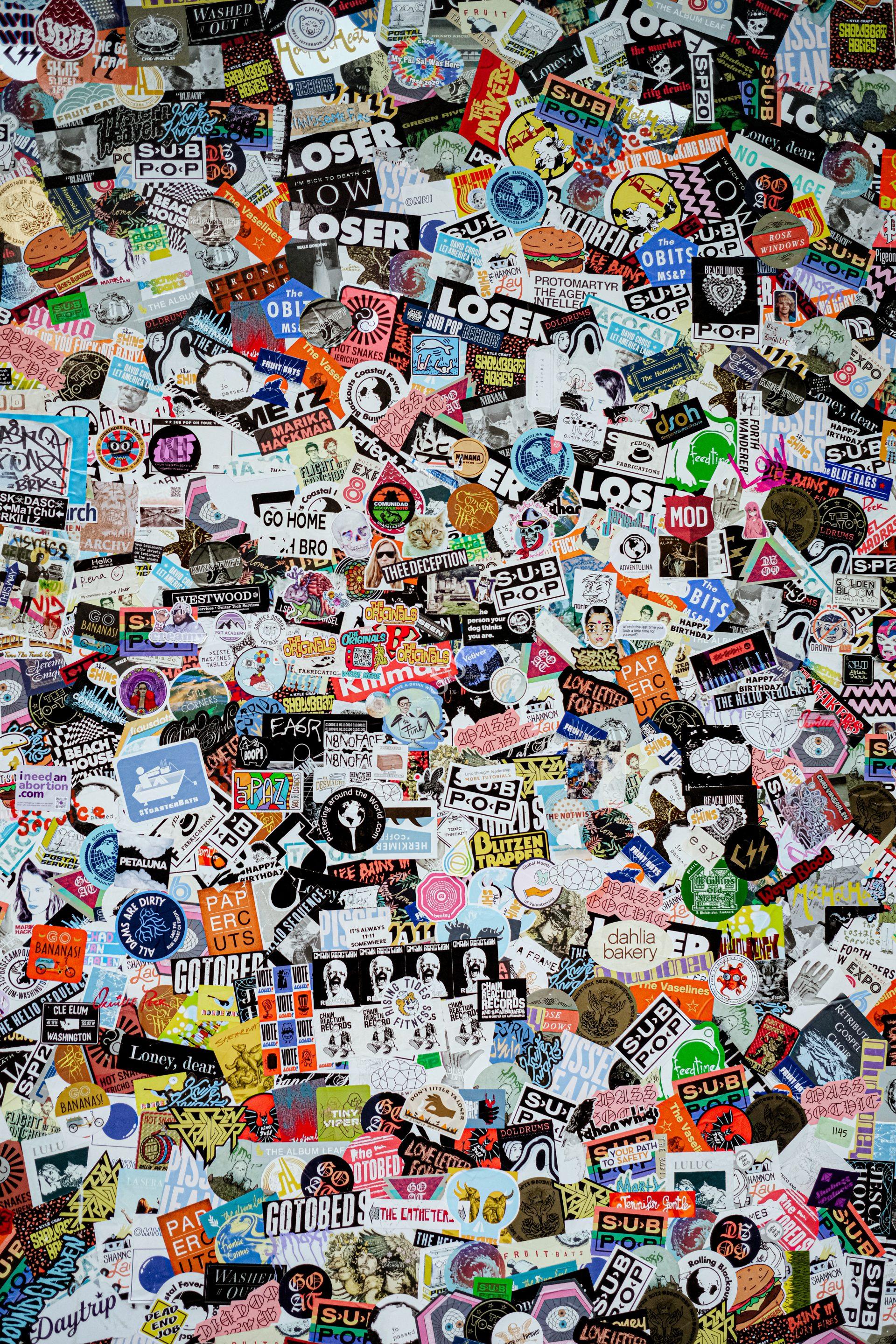 A large, colorful wall densely covered in a wide variety of stickers featuring slogans, logos, drawings, and cartoon characters, overlapping each other and creating a chaotic, vibrant collage.