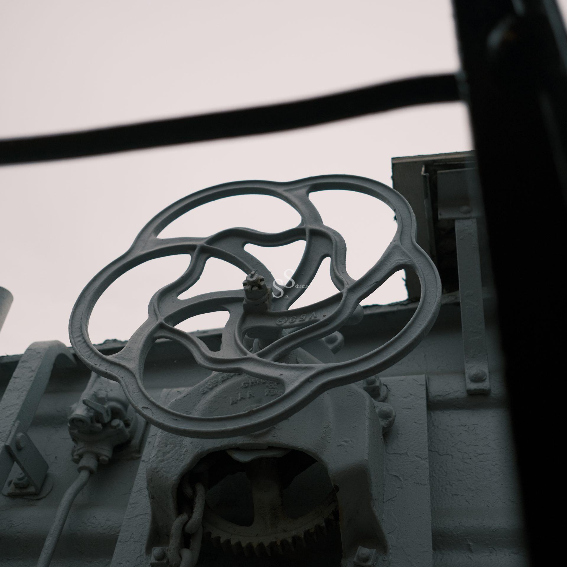 A close-up, low-angle view of a large, industrial metal valve wheel attached to train machinery, with an overcast sky in the background.