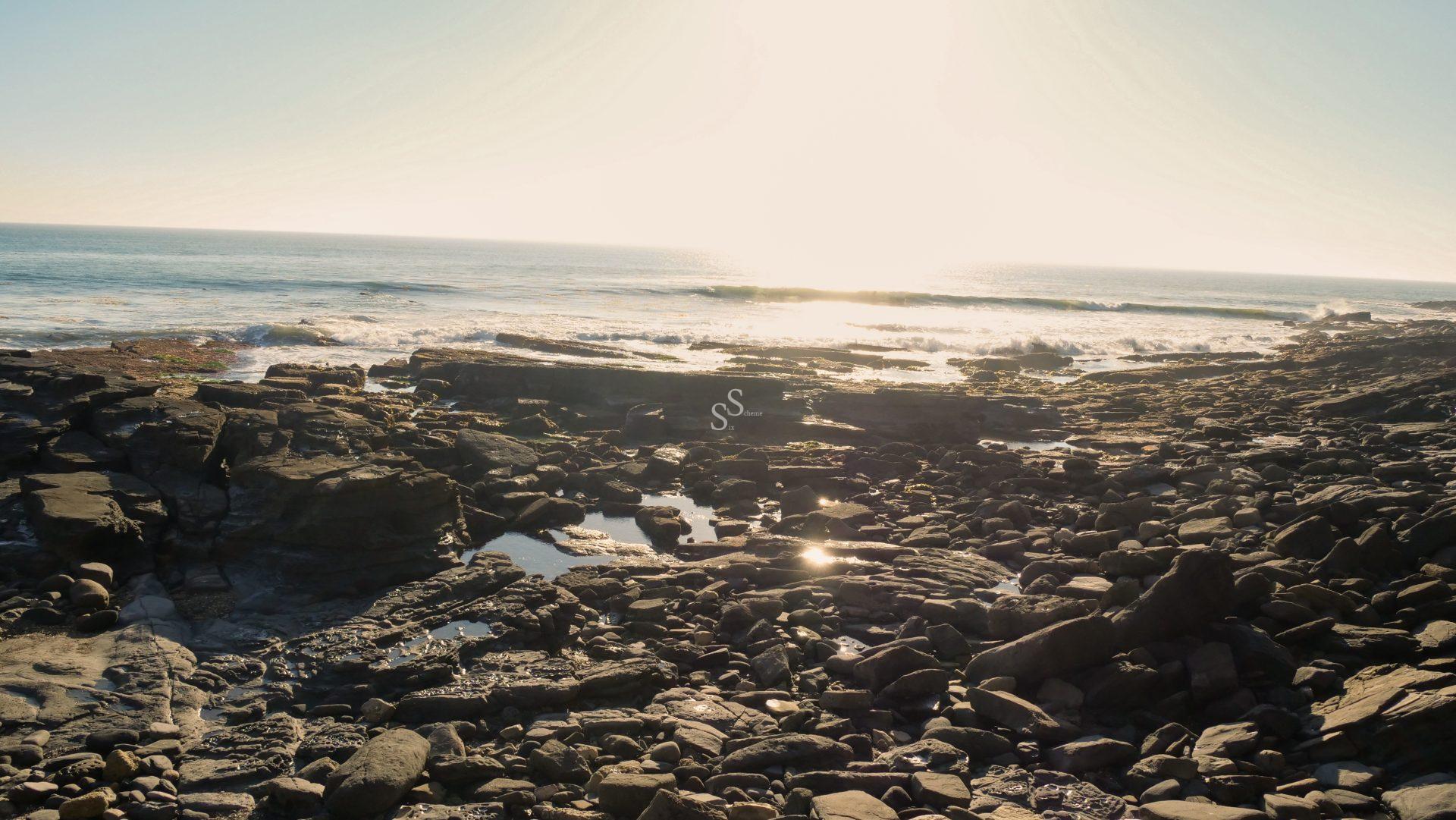 Rocky coastline with scattered tidal pools under bright sunlight, waves gently crashing in the distance, and the sun low on the horizon, creating a warm, glowing atmosphere over the ocean.