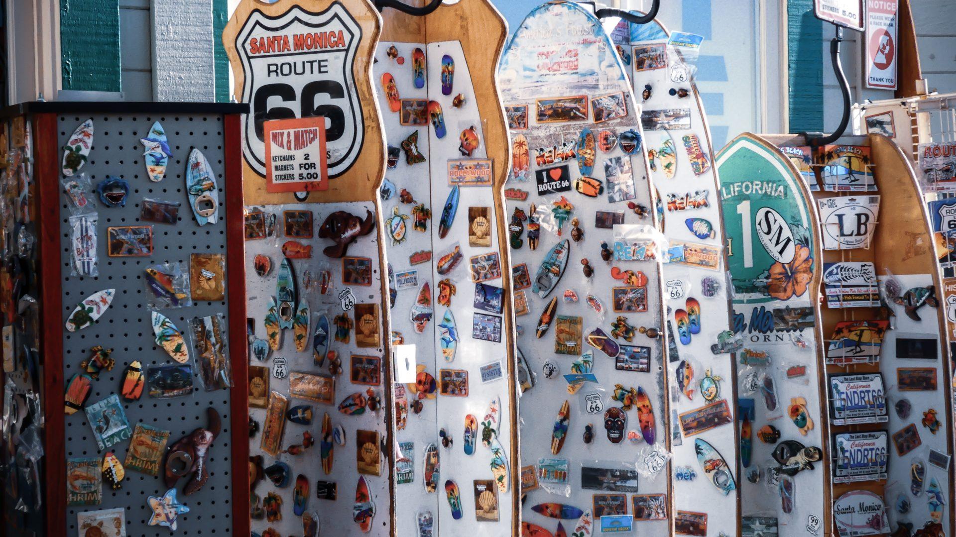 A display of colorful magnets and souvenirs on surfboard-shaped stands, featuring Route 66 signs, California and beach-themed images, and postcards, set up outdoors in a sunny location.