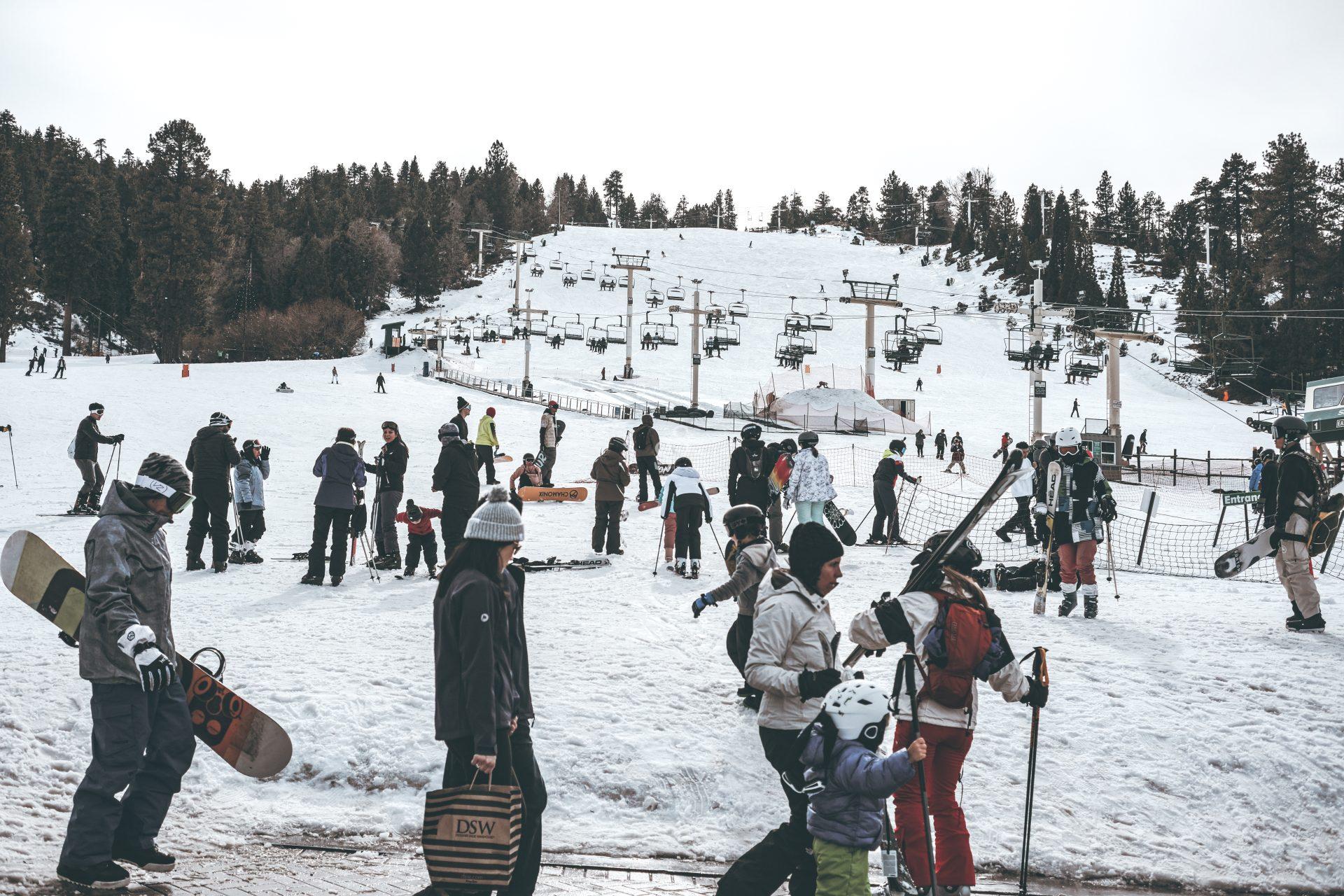 People in winter clothing gather at a snowy ski resort, some carrying skis and snowboards. Ski lifts and skiers are visible on the slopes in the background, with trees lining the edges of the scene.