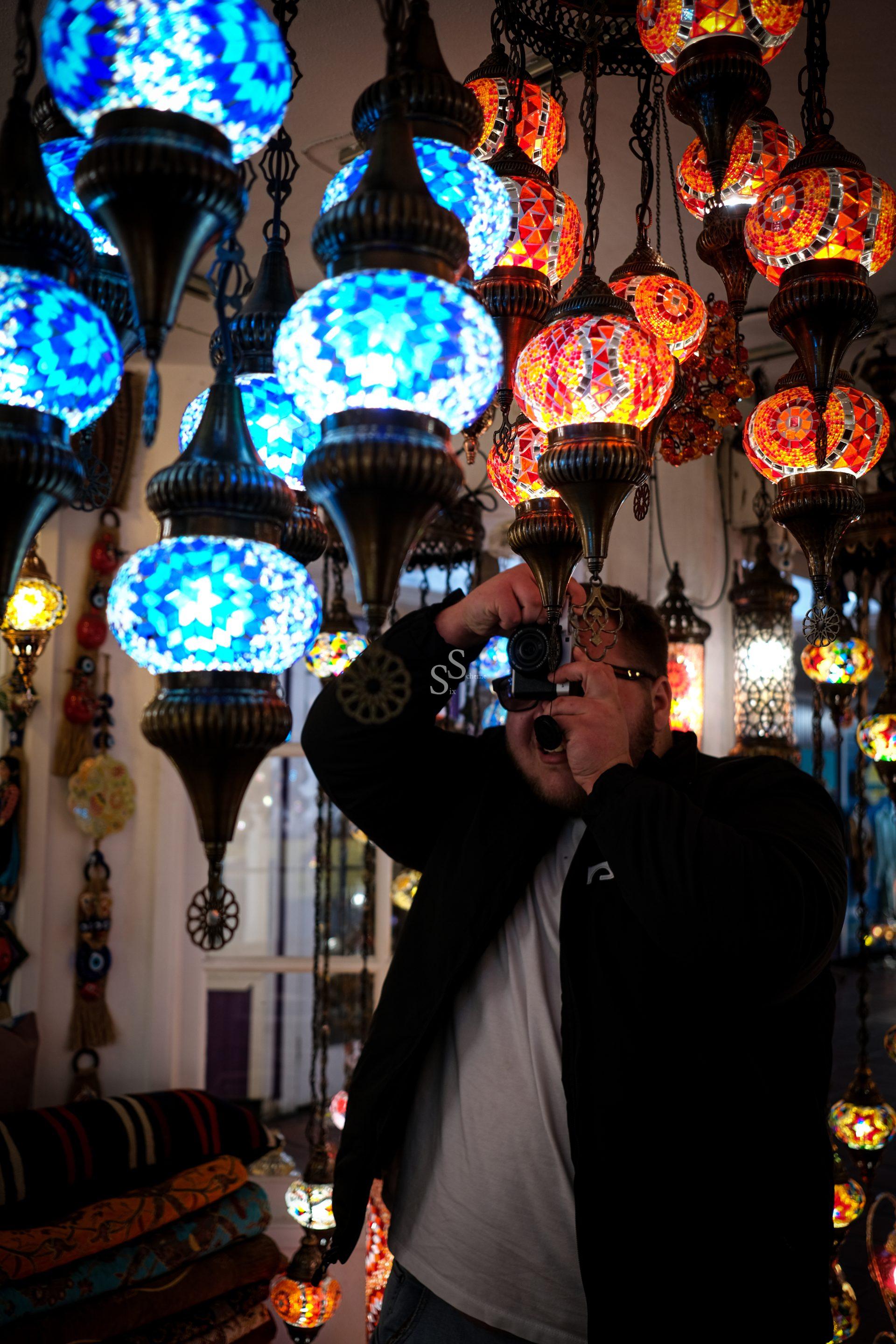 A person holding a camera takes a photo amid a vibrant display of colorful, mosaic-patterned hanging lamps in blue, red, and orange tones, creating an artistic and lively atmosphere.