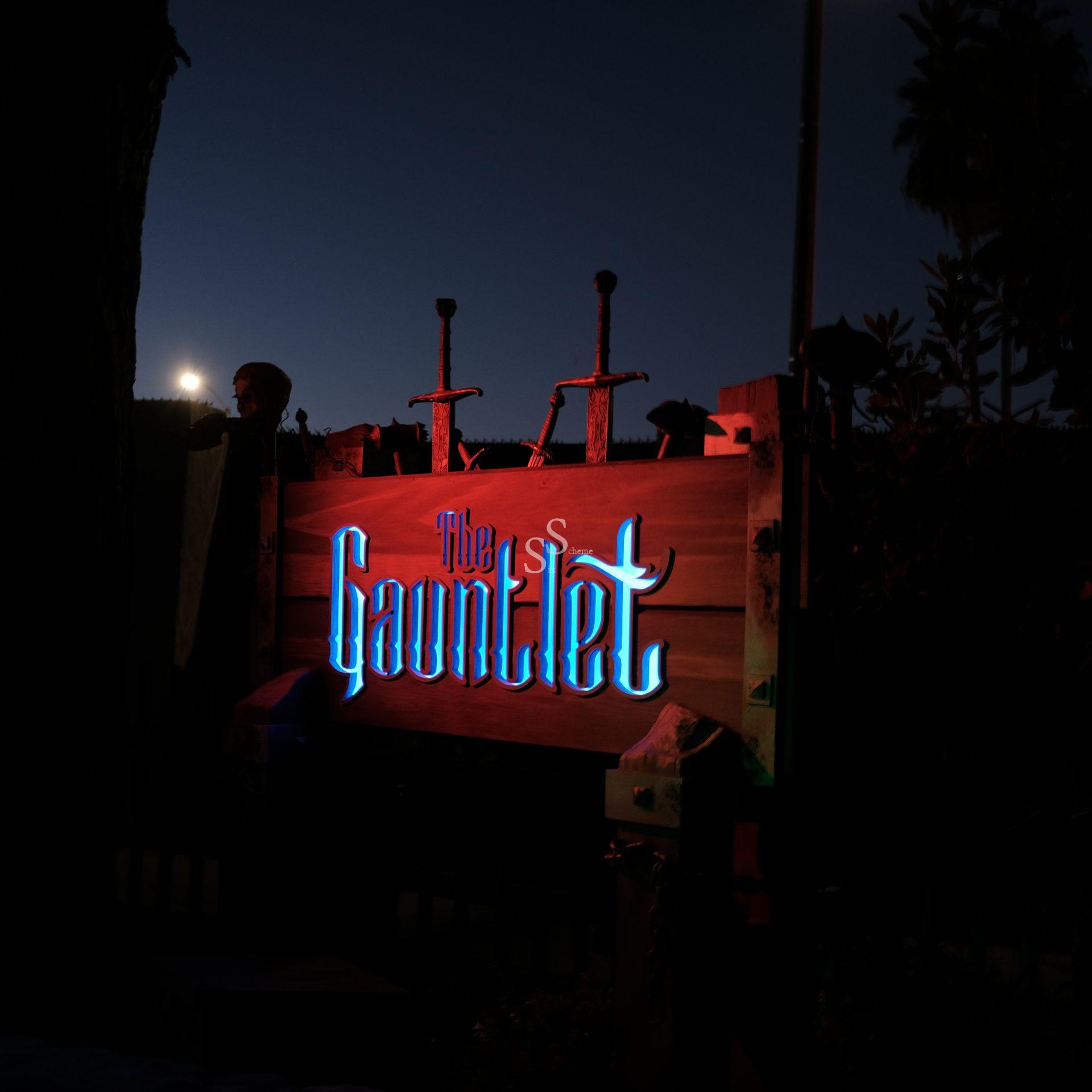 A wooden sign reading The Gauntlet in glowing blue letters is illuminated at night, with three swords sticking out from behind and trees silhouetted in the background.