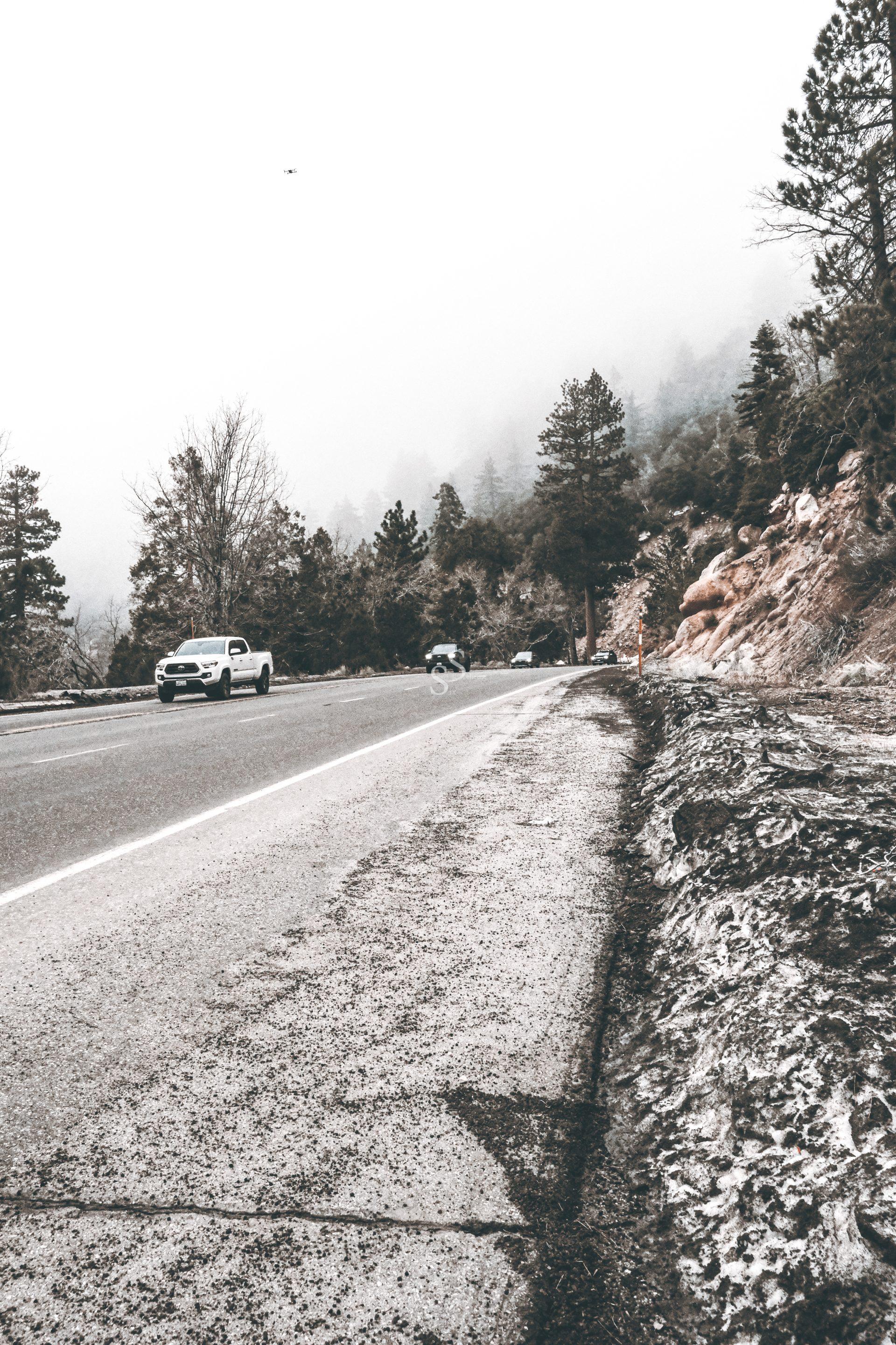 A misty, tree-lined mountain road with a few cars parked along the side and patches of snow on the ground; the scene has a cold, overcast atmosphere.