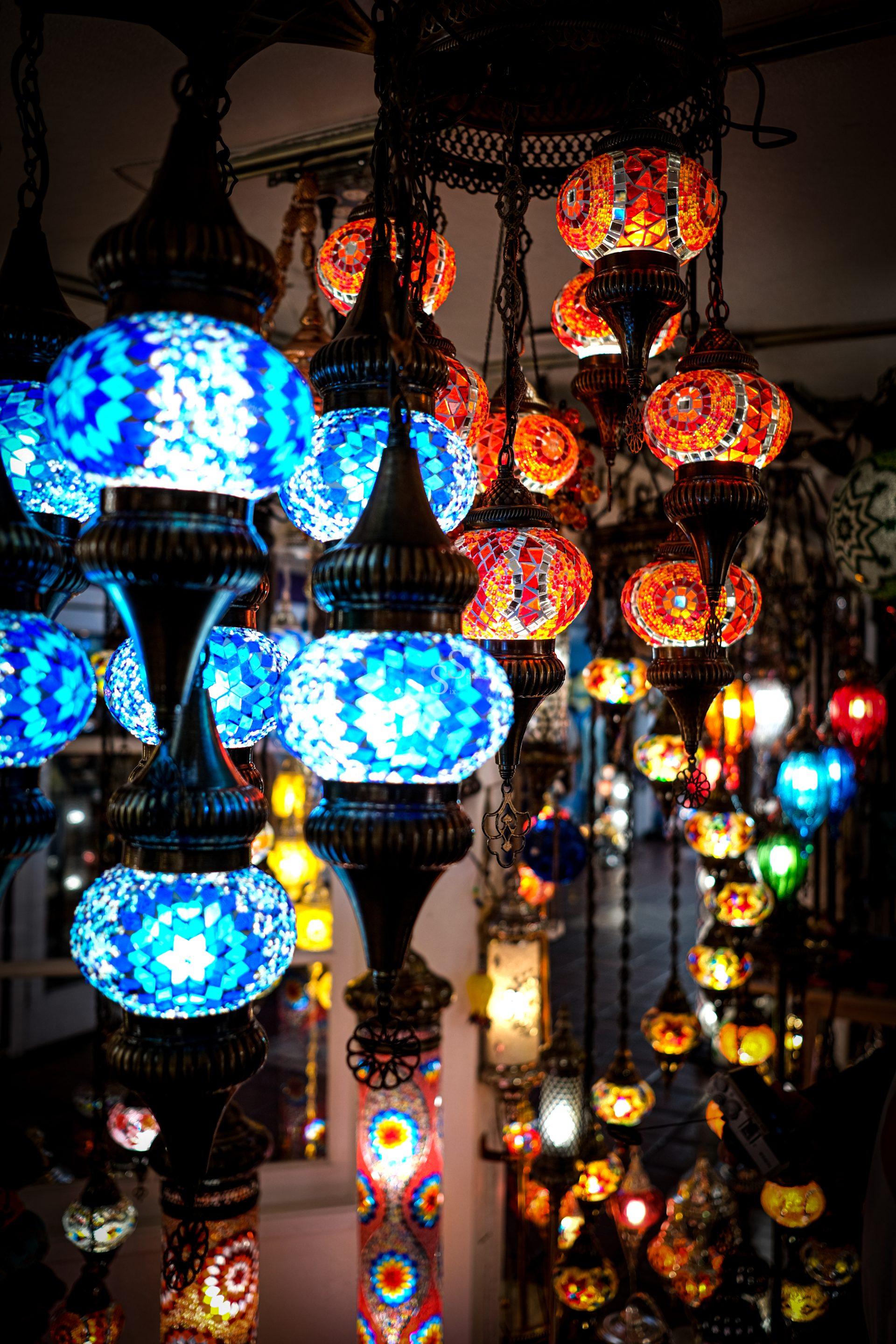 Colorful mosaic lamps hang from the ceiling, glowing in shades of blue, red, orange, and yellow, creating a vibrant and intricate display in what appears to be a market or shop setting.