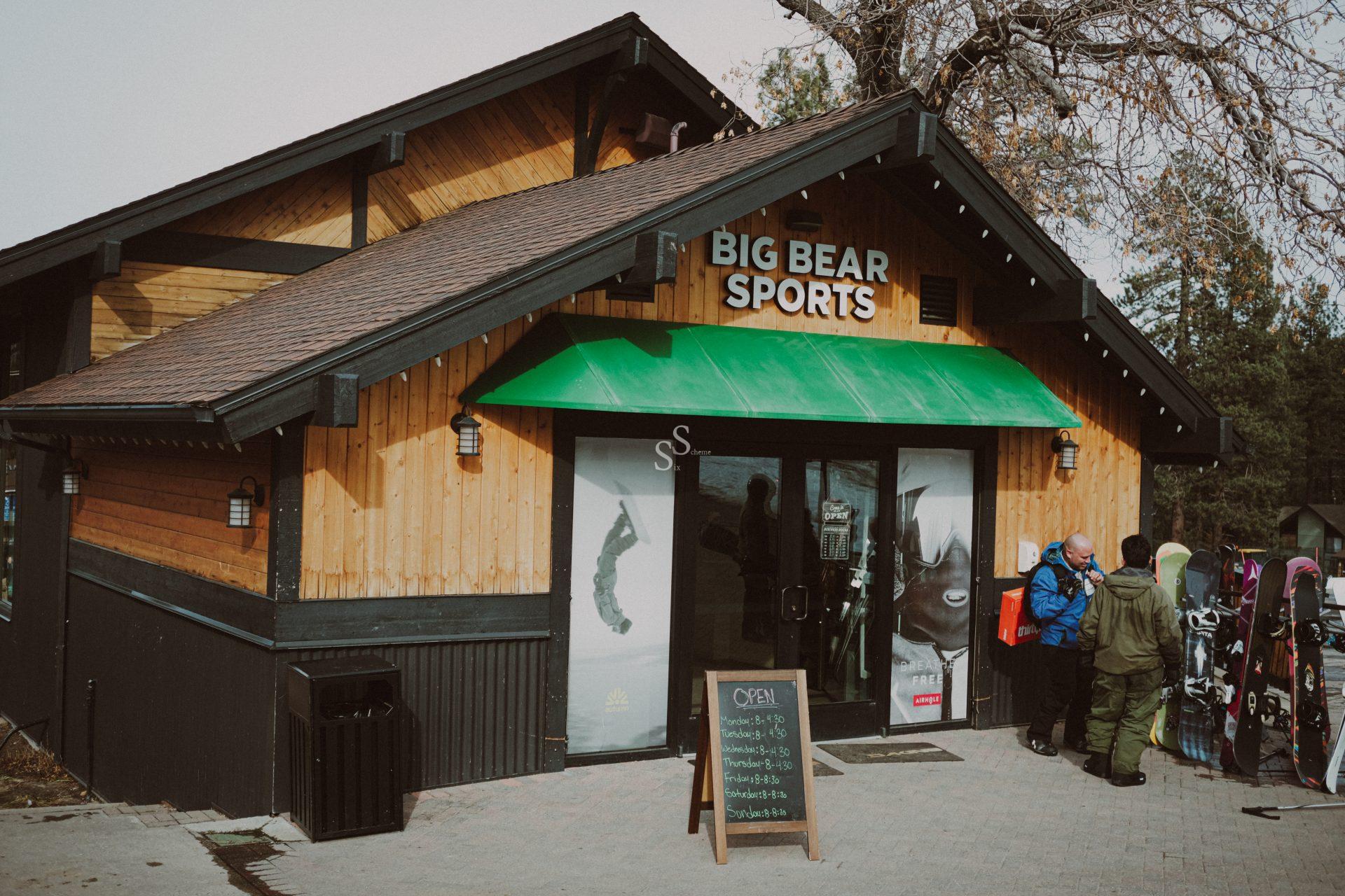 A wooden building labeled Big Bear Sports has a green awning above the entrance. Several people in winter clothing and ski gear stand outside near a chalkboard sign and racks of snowboards or skis.