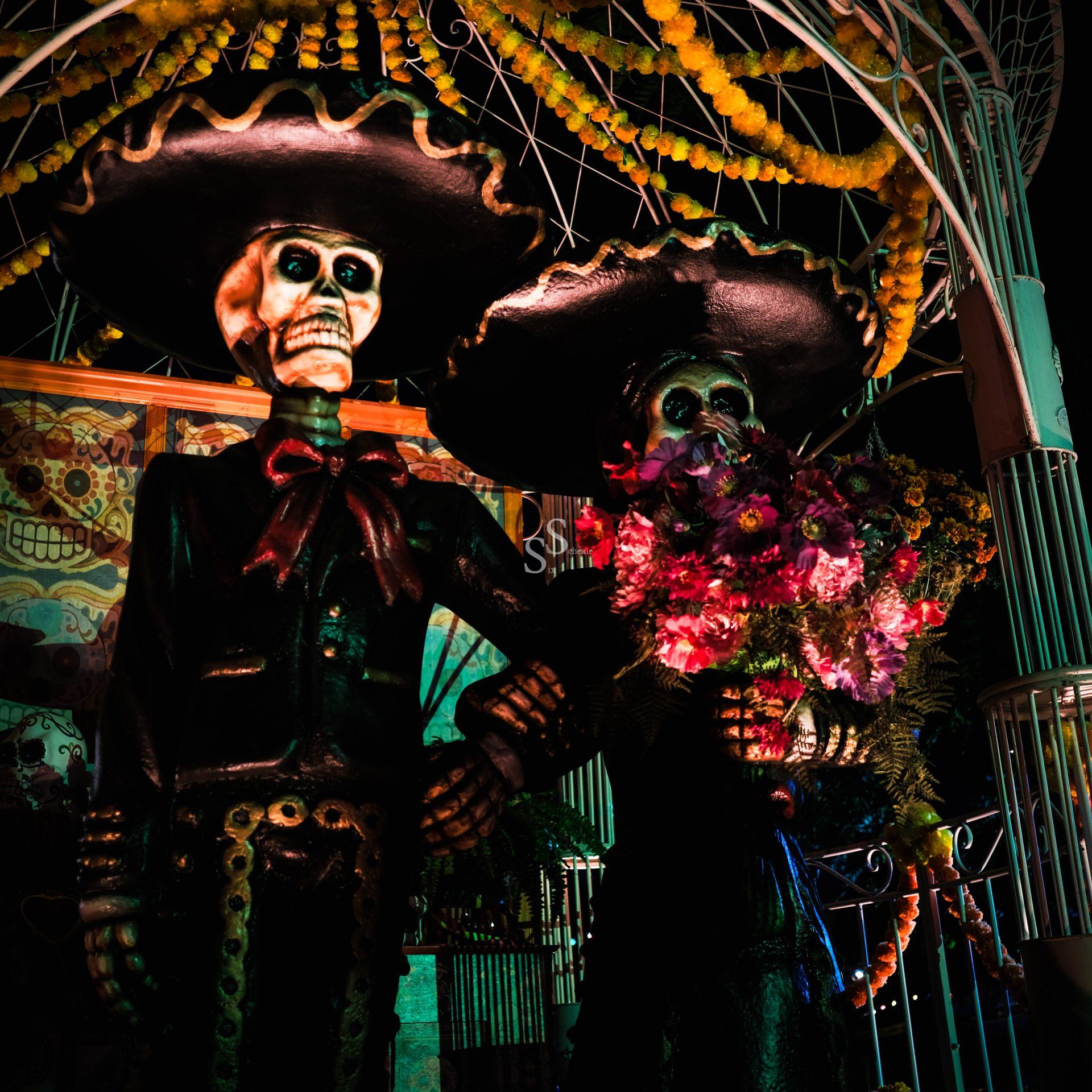 Two large skeletal figures dressed in traditional Mexican clothing stand arm in arm, one holding a bouquet of colorful flowers, under festive yellow decorations, celebrating Día de los Muertos at night.