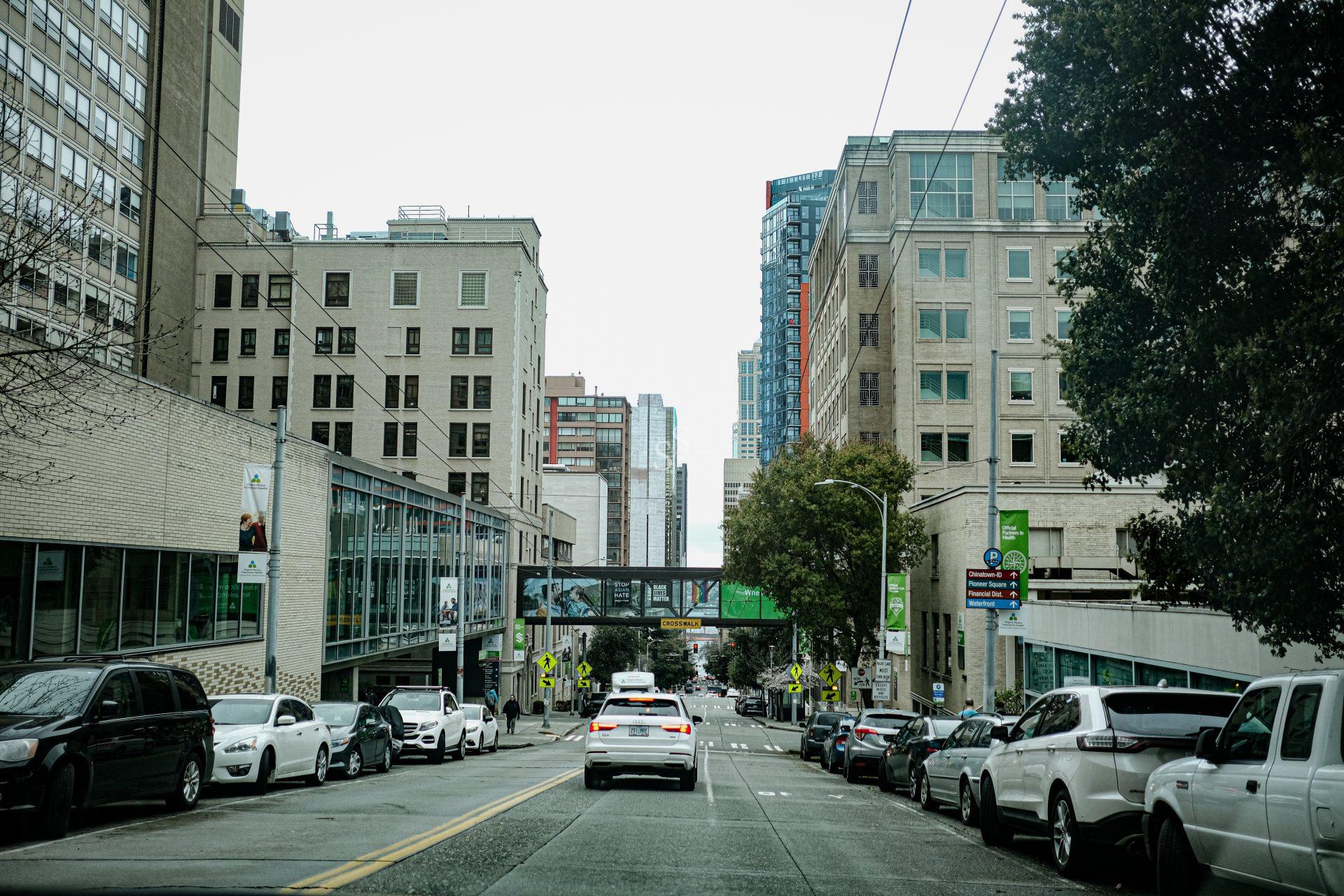 A city street lined with parked cars on both sides, tall buildings, and a skybridge connecting two structures. A white car drives down the road toward the background. Trees and street signs are visible.