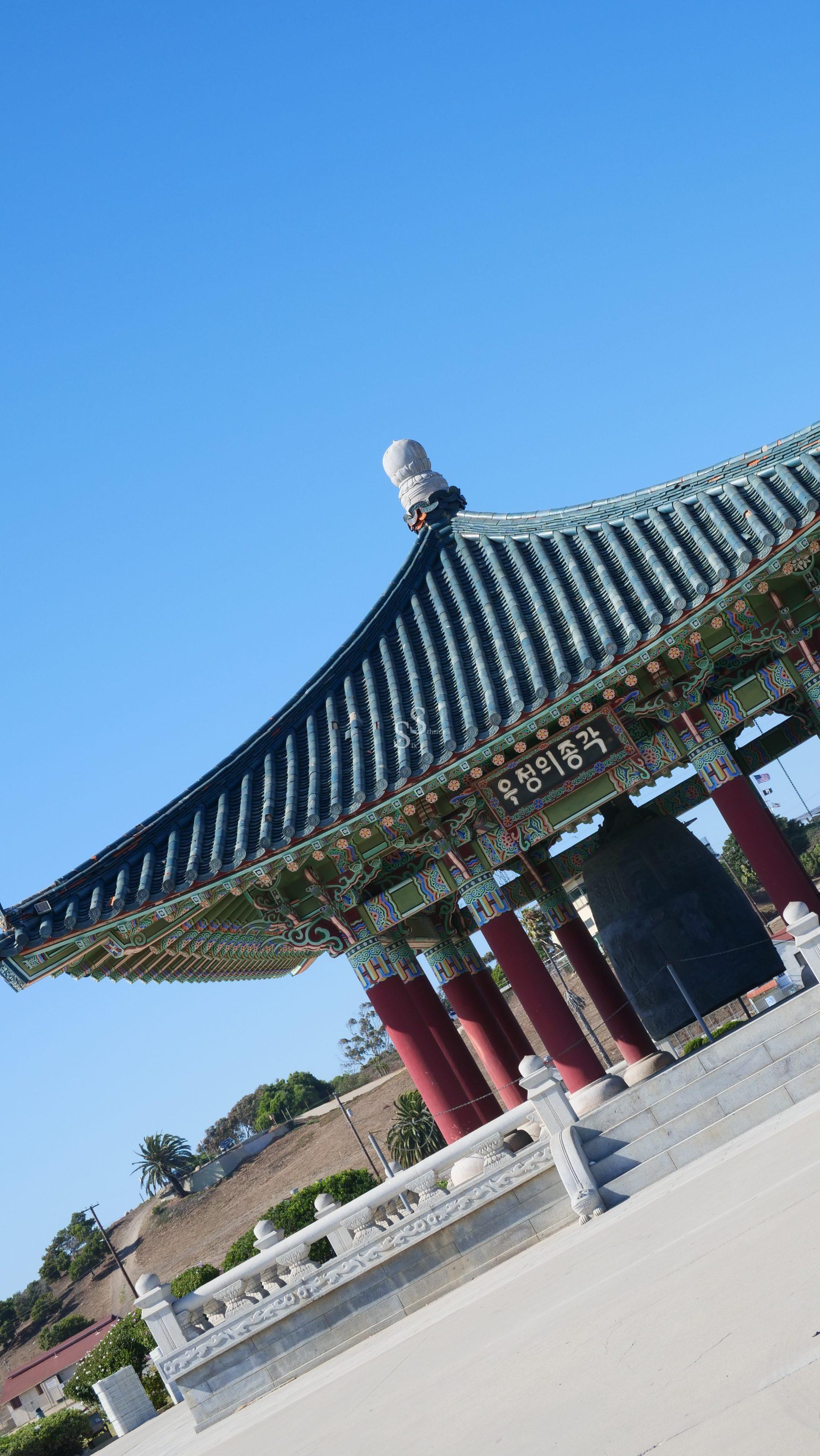 A traditional Korean pagoda-style pavilion with ornate painted designs and a large hanging bell stands under a clear blue sky, surrounded by stone steps and greenery.