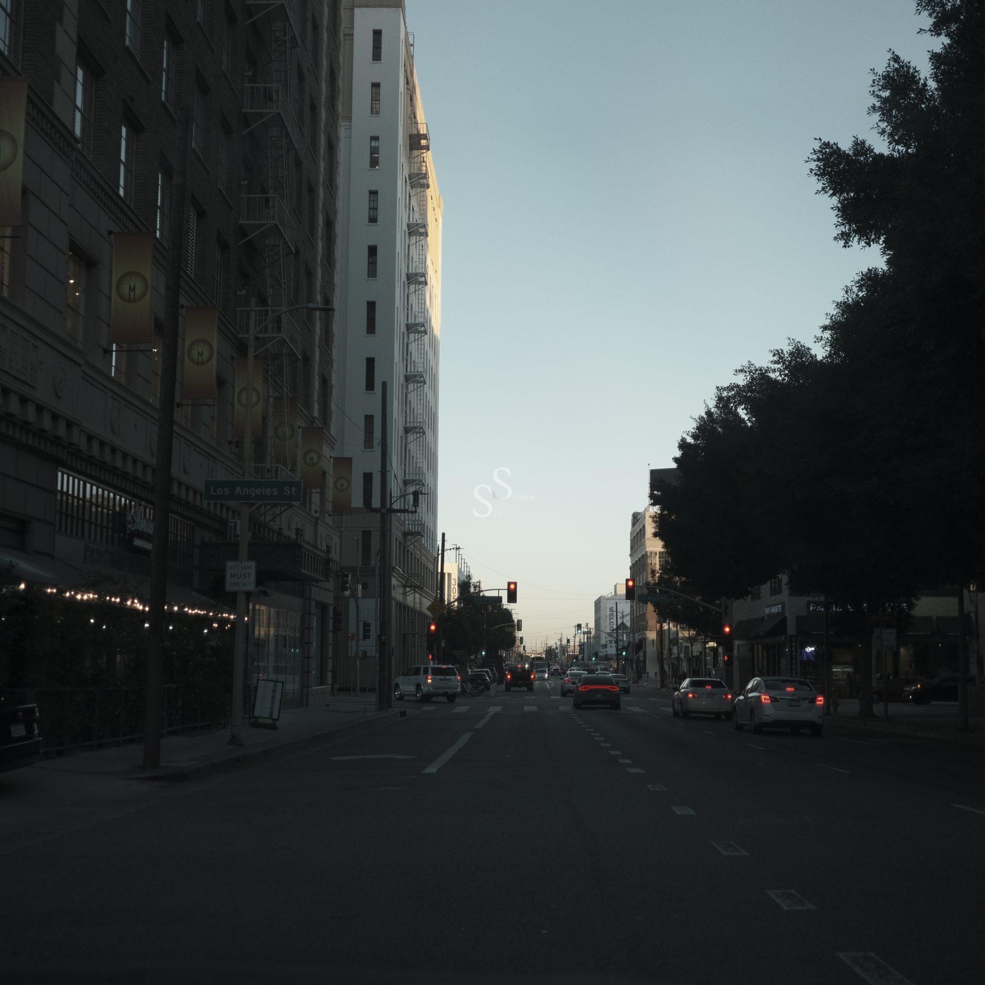 A city street at dusk with cars stopped at a red light. Tall buildings line both sides, and sunlight illuminates one building. Trees partially shade the right side, and street signs are visible on the left.