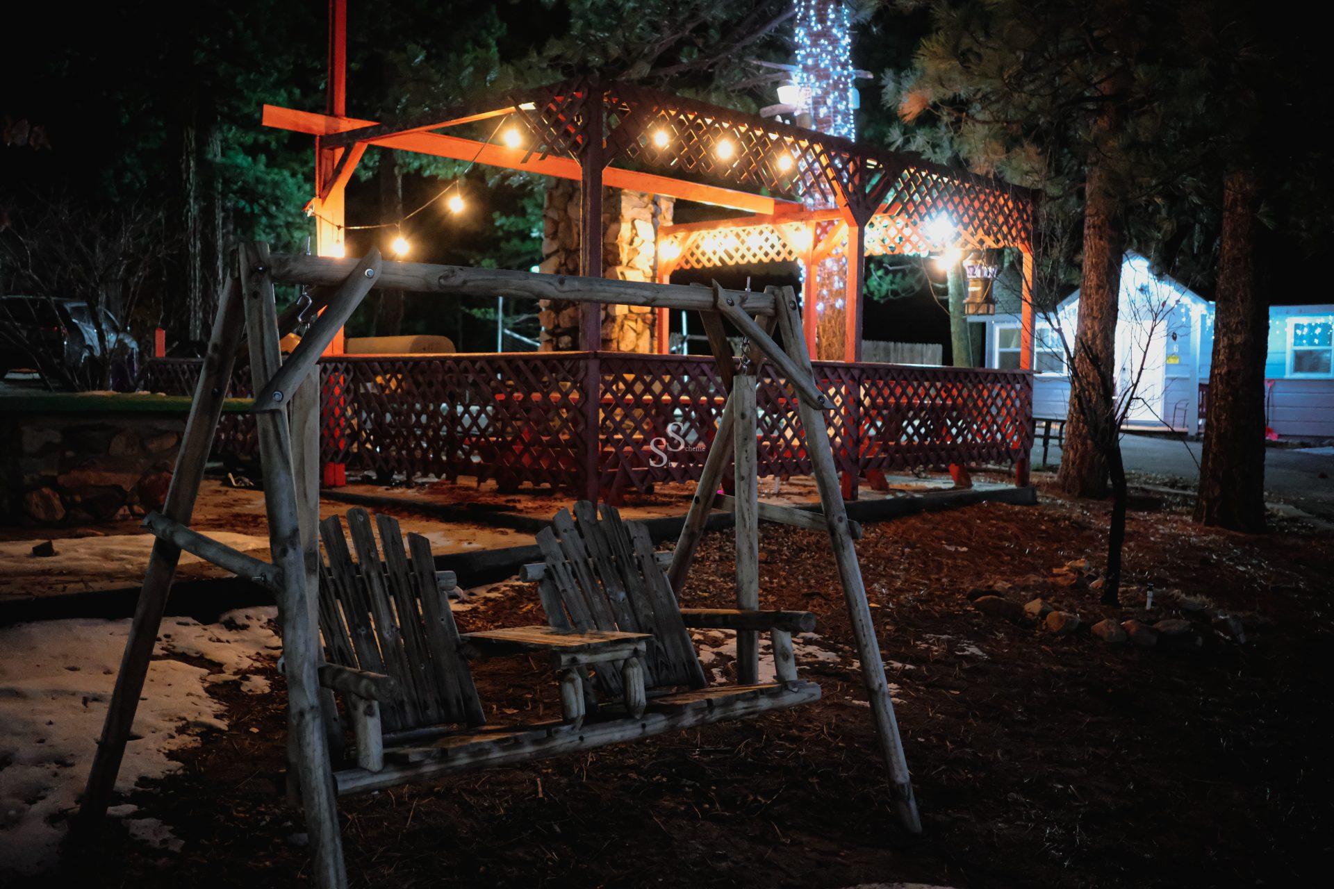 A rustic wooden swing for two sits in the foreground, with a warmly lit outdoor patio, string lights, and trees in the background at night. Some snow is on the ground.