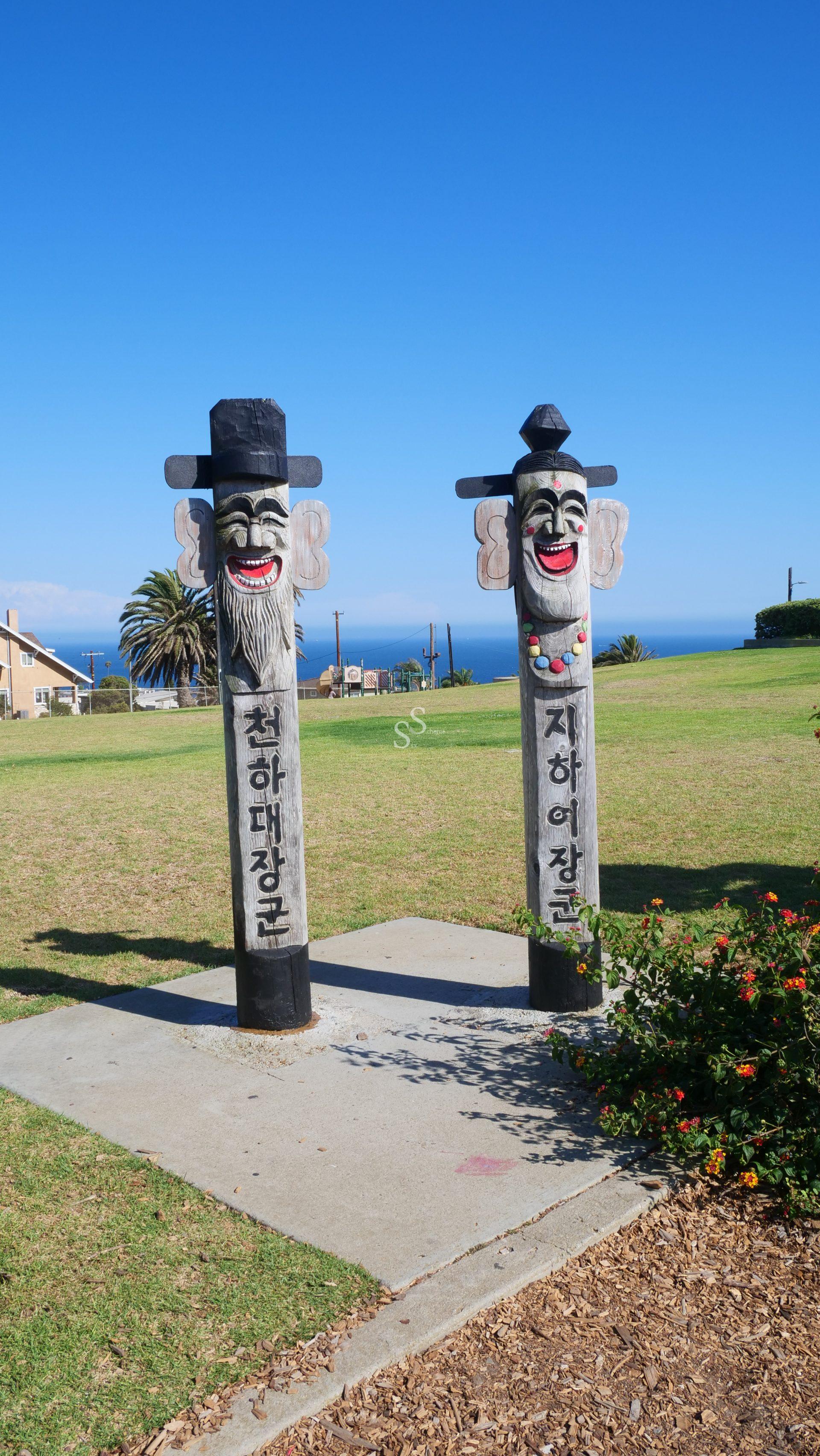Two tall, wooden Korean totem poles with painted faces stand on a grassy park overlooking the ocean, under a clear blue sky. Shrubs and a building are visible in the background.