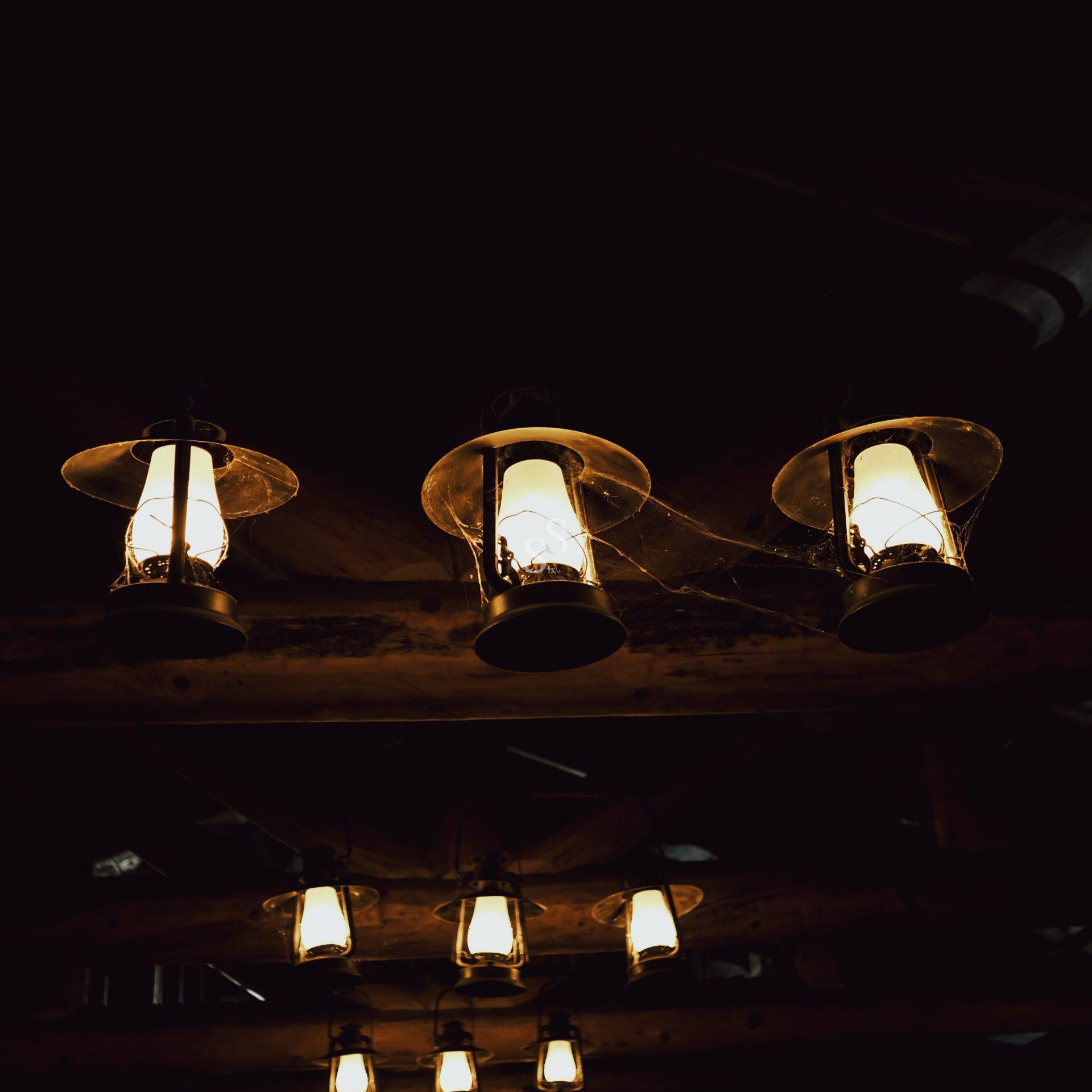 A row of old-fashioned lanterns hang from a dark wooden ceiling, casting a warm, soft light. Some lanterns have visible cobwebs, creating a rustic and slightly eerie atmosphere.