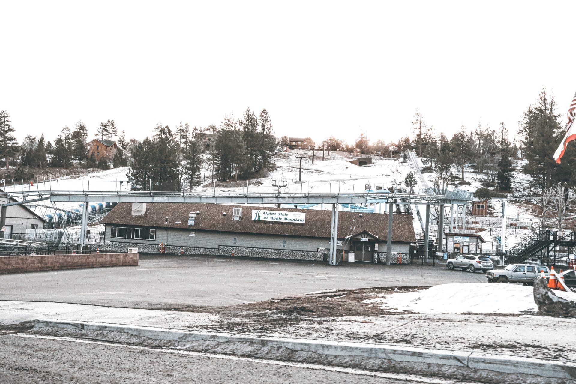 A ski resort with a main lodge building, ski lifts, and patches of snow on the slopes and ground. Several parked cars are visible, along with trees and houses in the background under an overcast sky.
