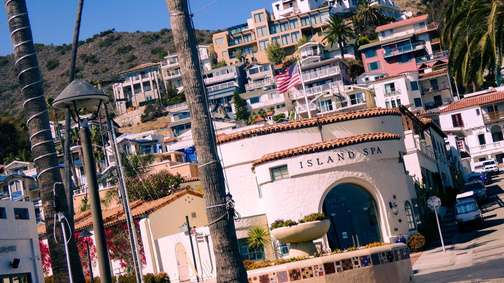 Mediterranean-style buildings on a hillside overlook a white stucco spa with an arched entrance and an American flag; palm trees and a tiled fountain are visible in the foreground under a clear blue sky.