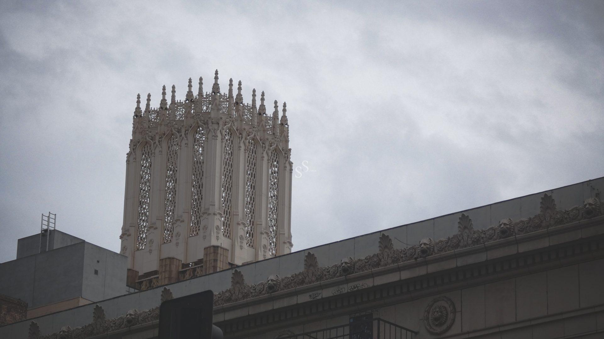 An ornate, white Gothic-style tower rises above a decorated building facade under a cloudy, gray sky. The tower features intricate architectural detailing and pointed spires.