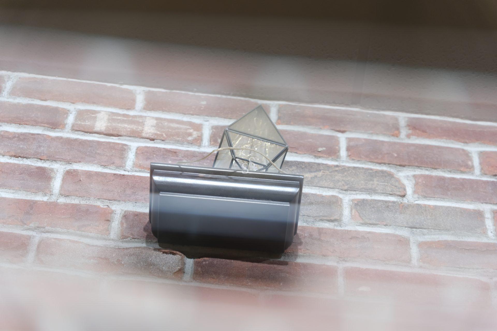 A wall-mounted outdoor light fixture with a geometric glass shade is attached to a red brick wall, viewed from below at an angle.