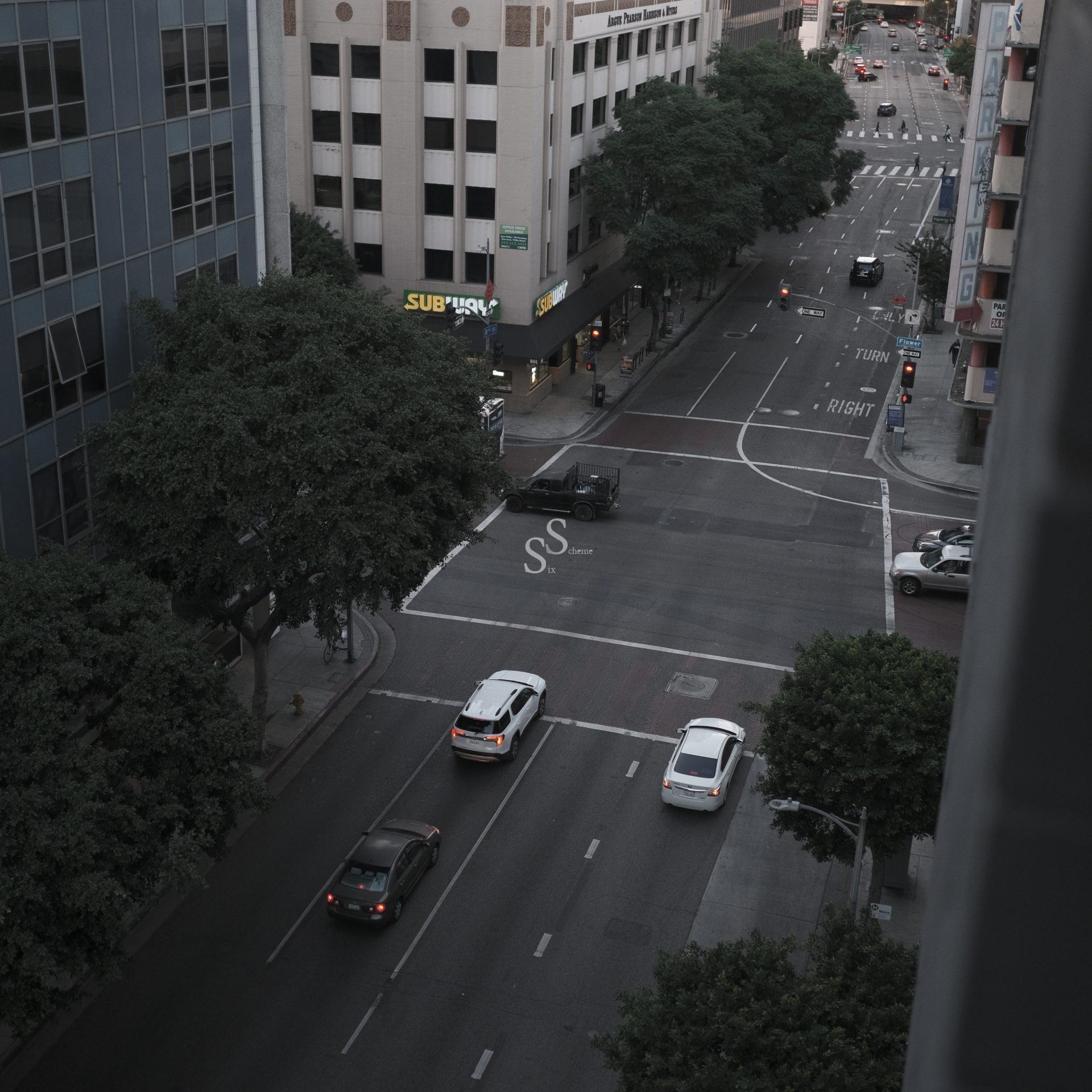 A city street intersection viewed from above, with cars stopped at a red light. Trees line the sidewalks, and buildings, including a Subway restaurant, are visible. The scene appears quiet and overcast.