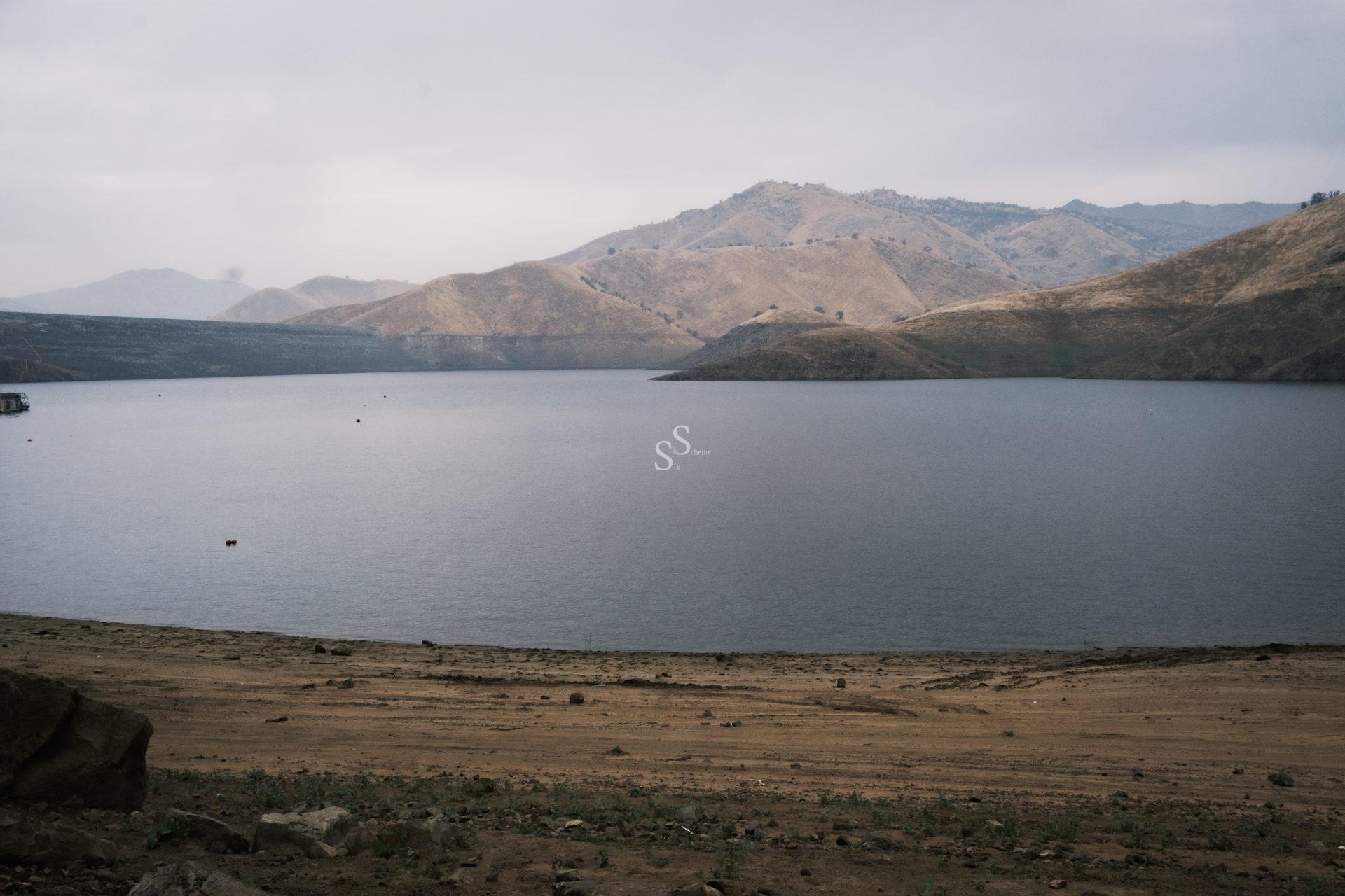 A calm lake bordered by a dry, sandy shore with rolling, brown hills in the background under an overcast sky.