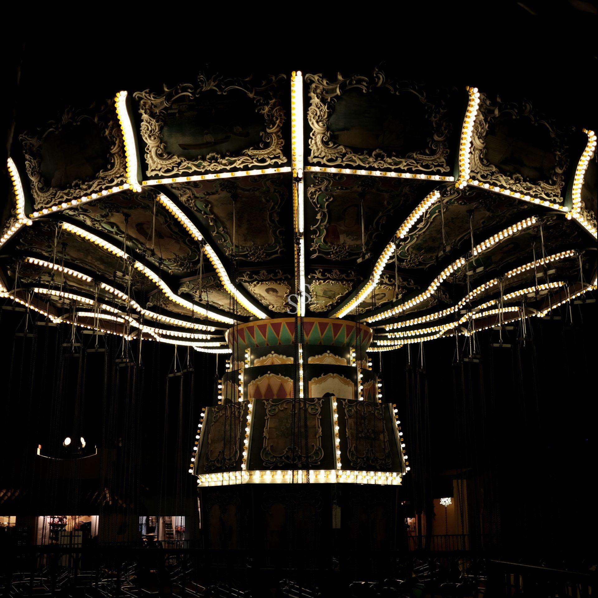 A brightly lit carousel swing ride glows against a dark night background, highlighting its ornate patterns and rows of empty hanging chains.