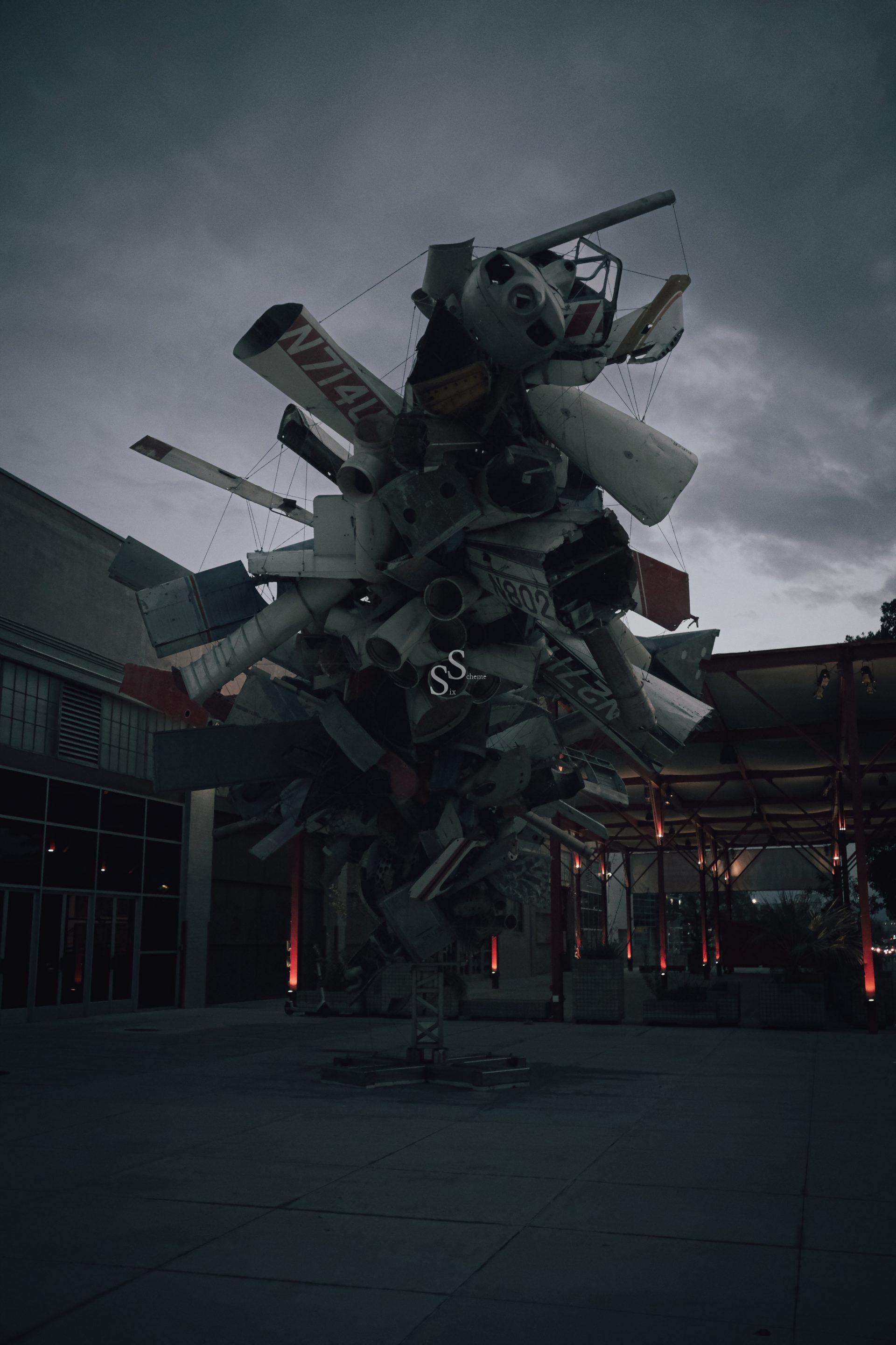 Crafted from airplane parts and metal, a large sculpture stands on a concrete plaza beside a building beneath a cloudy twilight sky.