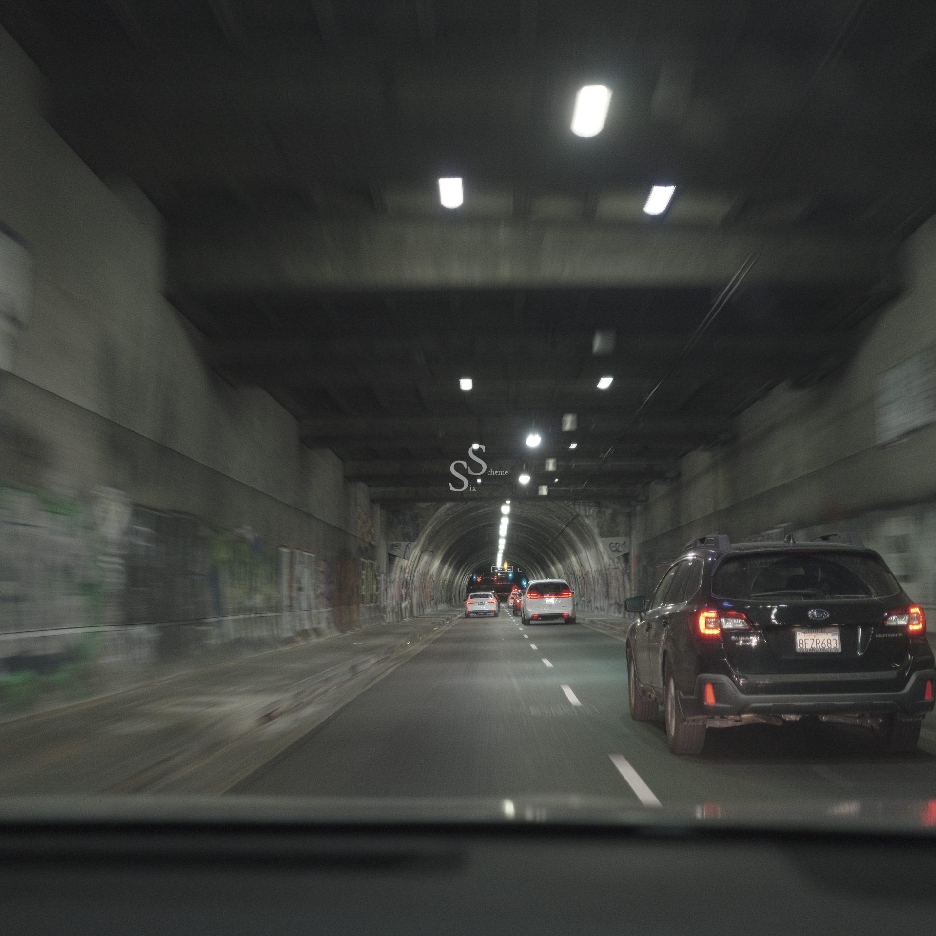 Cars drive through a dimly lit, graffiti-covered tunnel. The view is from inside another vehicle, showing blurred lights and walls as the road becomes slightly narrow ahead.
