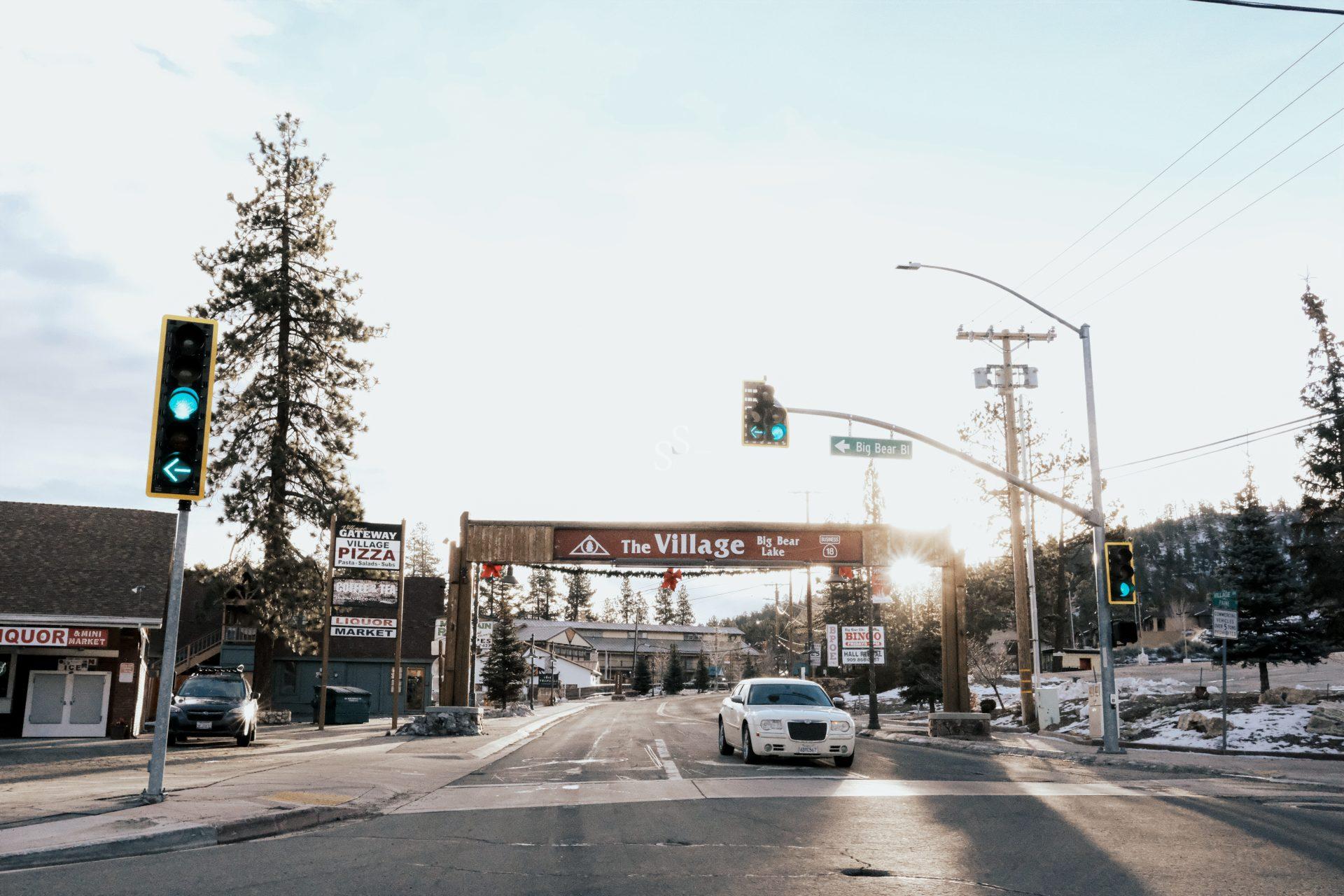 A car drives through an intersection toward a wooden archway reading The Village, Big Bear Lake on a sunny winter day, with green lights and some snow on the ground and trees.