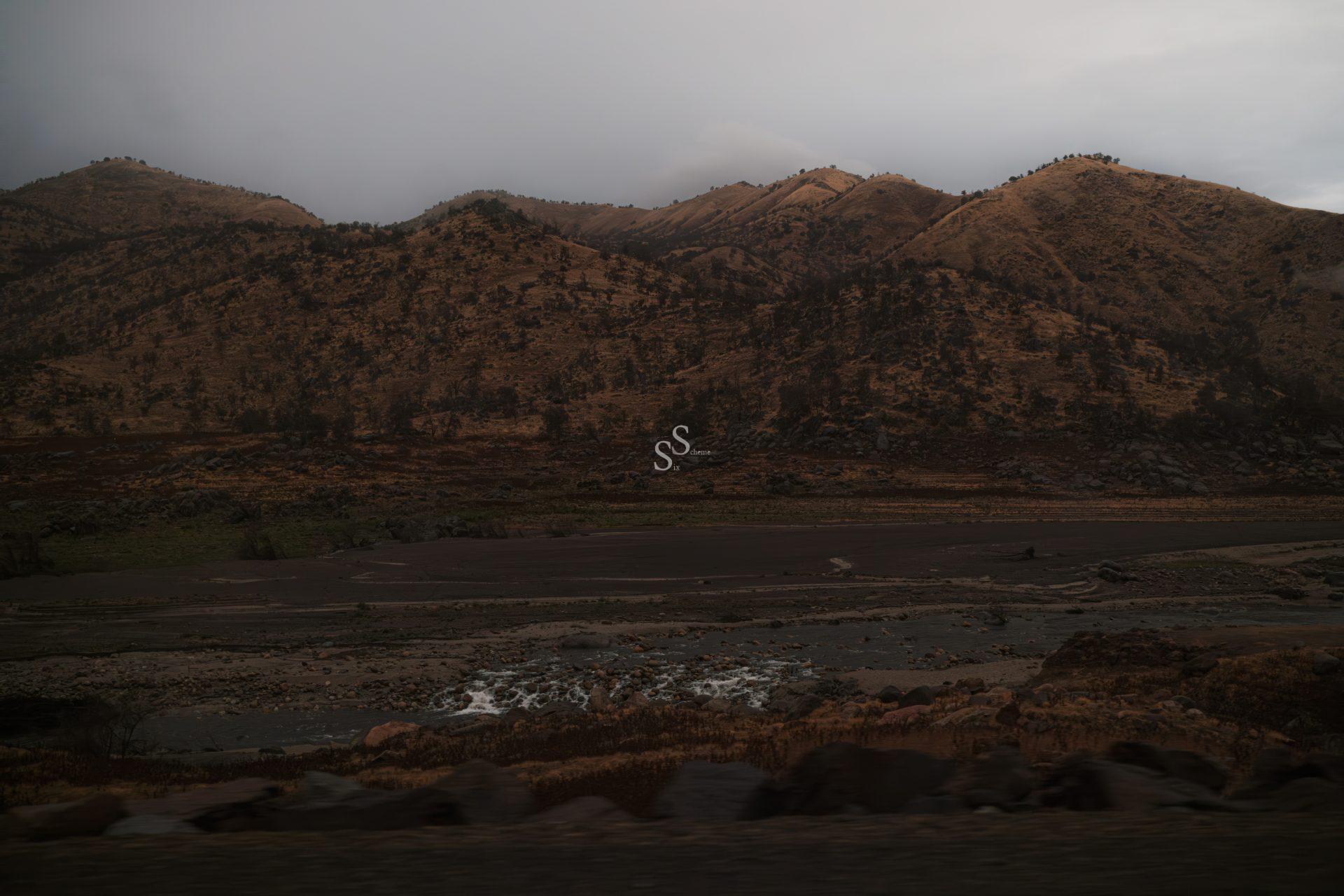 A cloudy sky over rugged, brown hills with sparse vegetation. In the foreground, a rocky riverbed with patches of flowing water moves through a dry, barren landscape.
