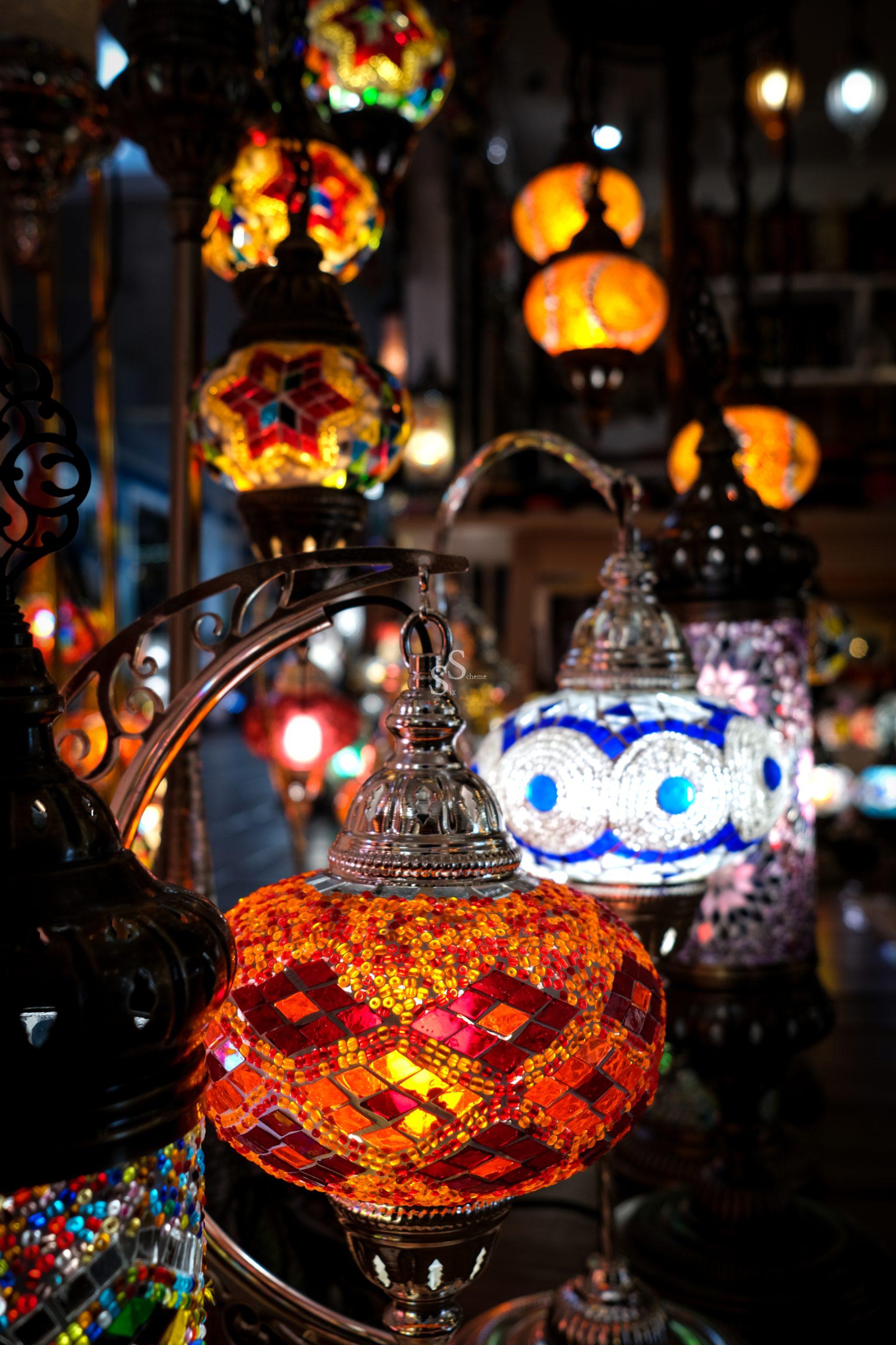Intricate blue and white patterns detail a mosaic lamp in close-up, set against a backdrop of other glowing, colorful lamps.