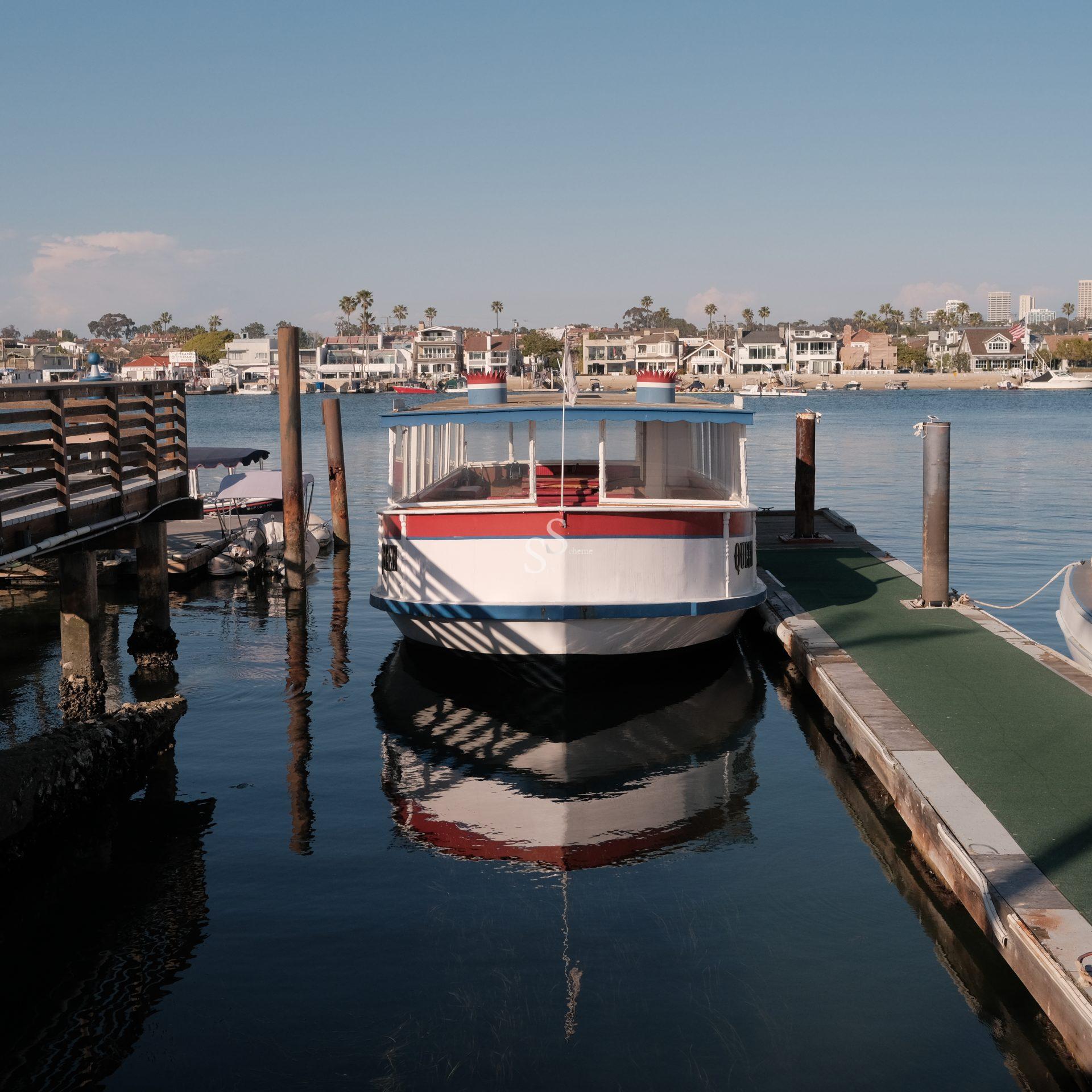A white boat with red trim is docked between wooden piers in a calm marina, with waterfront houses and palm trees visible in the background under a clear blue sky.