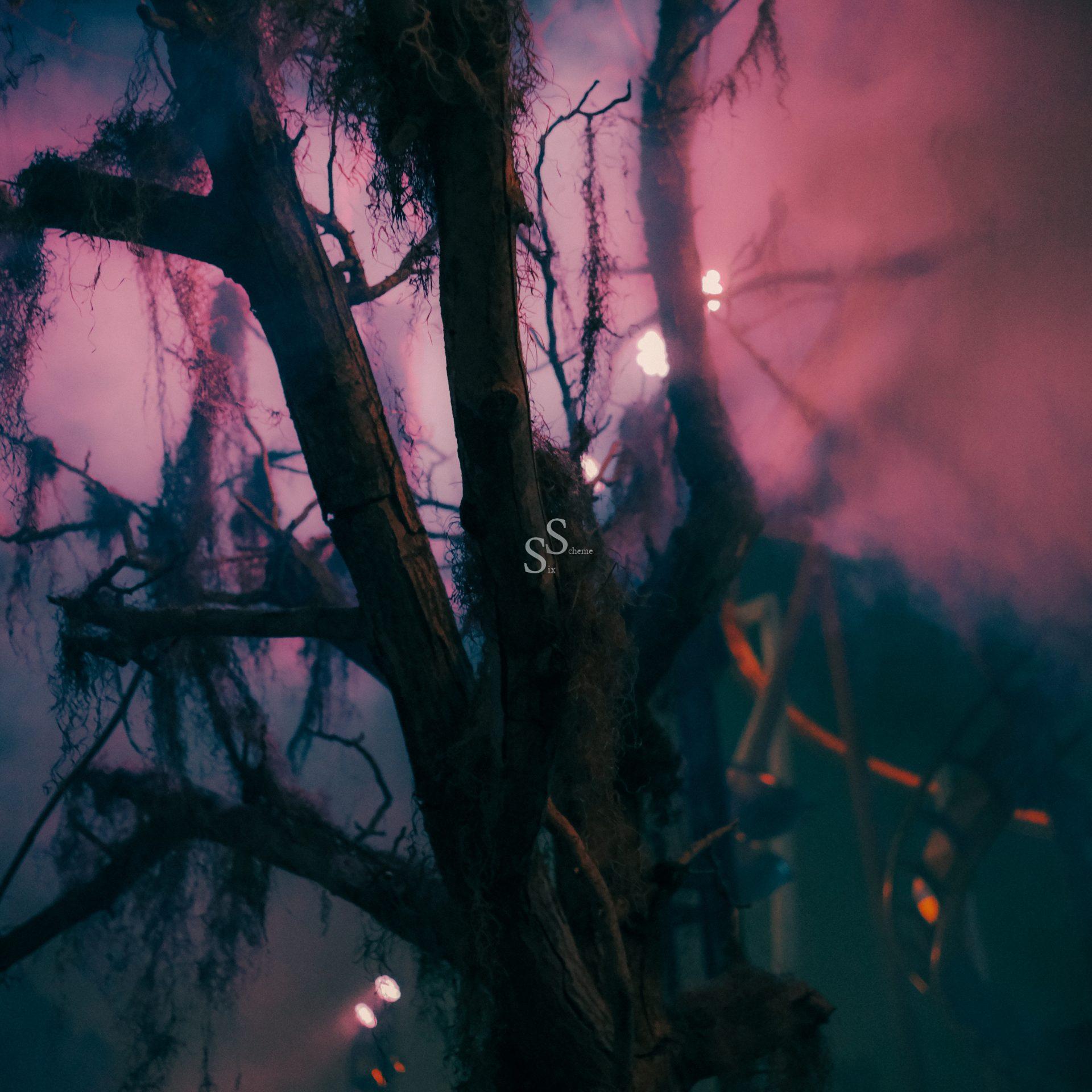 A dark, leafless tree with hanging moss stands in dense, colorful fog, illuminated by purple and blue light, creating a mysterious and eerie atmosphere.