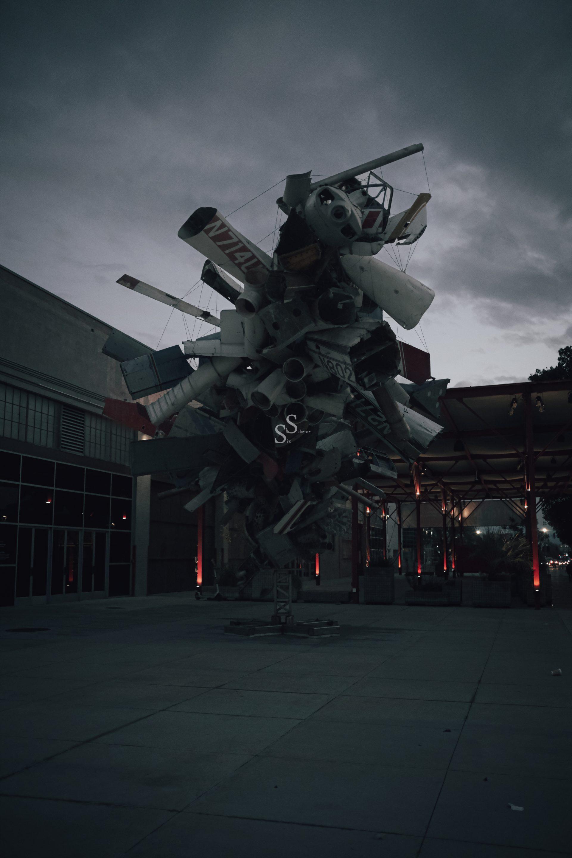 A large outdoor sculpture made of airplane parts and metal pieces stands on a concrete plaza near a building at dusk, beneath a cloudy sky.