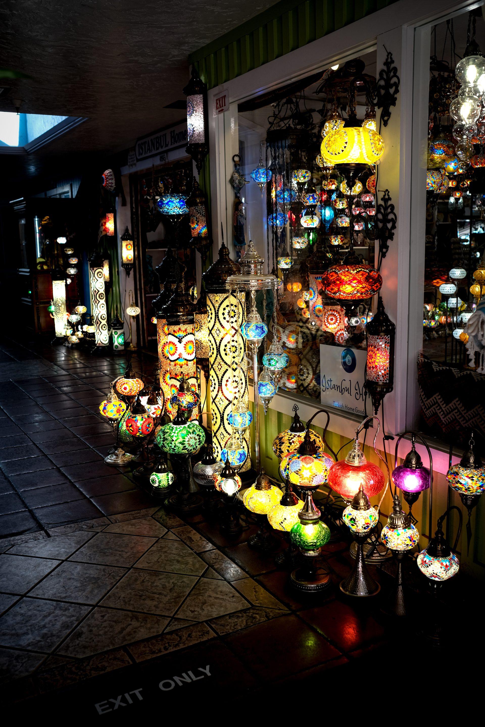 Colorful mosaic Turkish lamps, displayed on the floor and in windows of a dim shop, cast vibrant light onto the tiled floor.