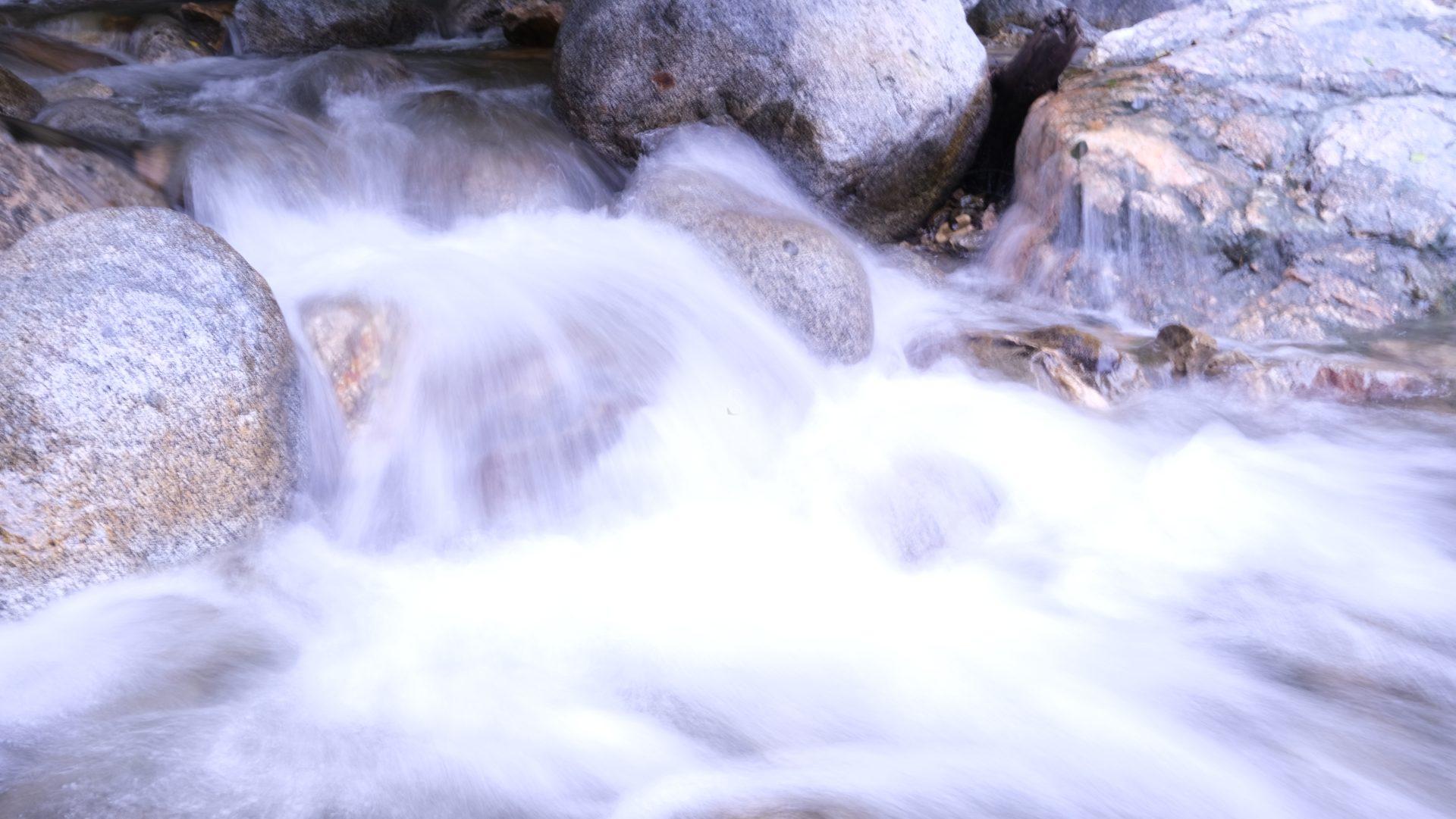 A close-up of a fast-flowing stream with white, rushing water cascading over smooth, large rocks. The water appears silky and blurred, suggesting a long exposure photograph.