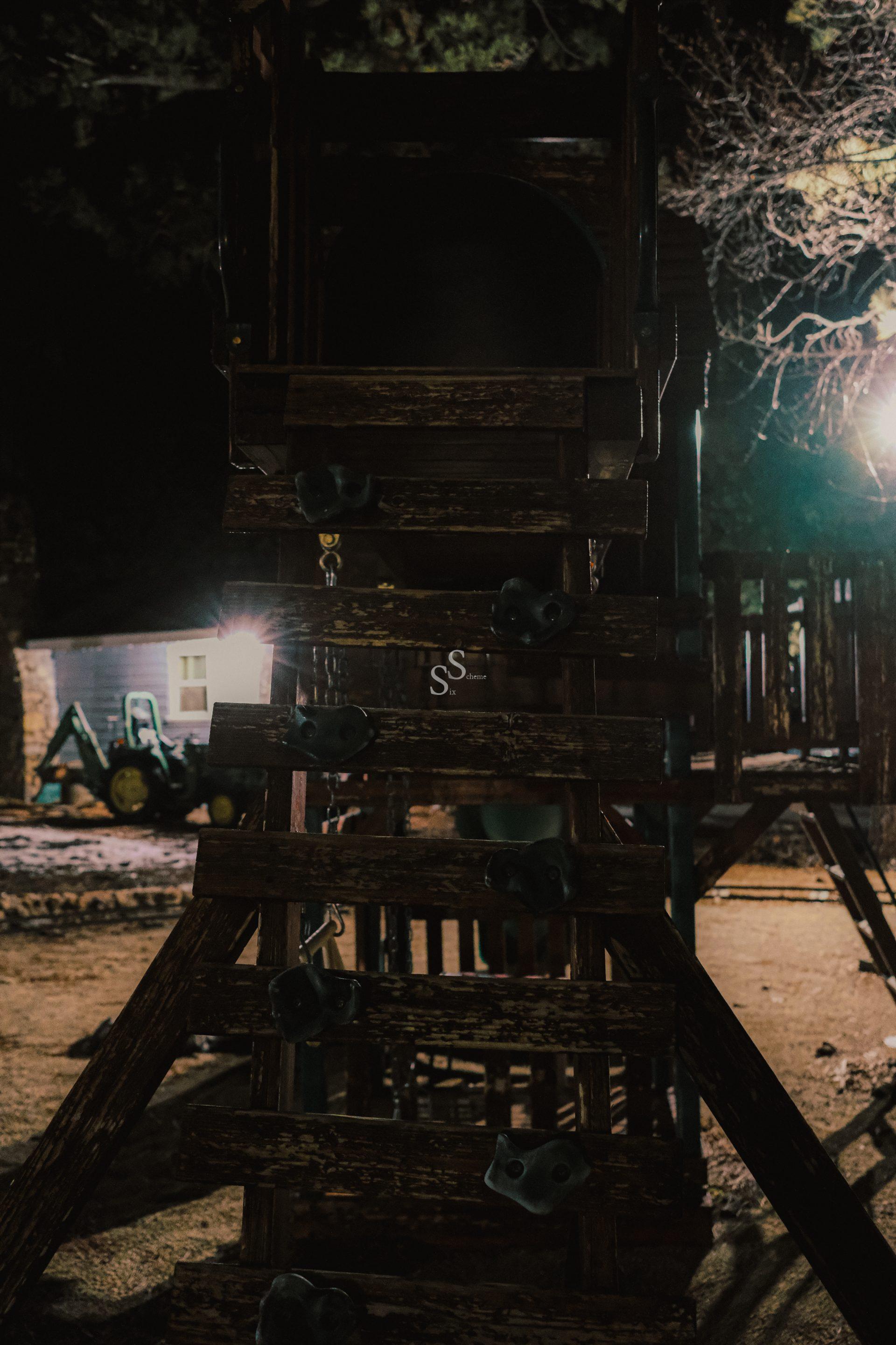 A dimly lit wooden playground climbing structure at night, with artificial lights and leafless trees visible in the background. A small green tractor is parked near a building on the left.