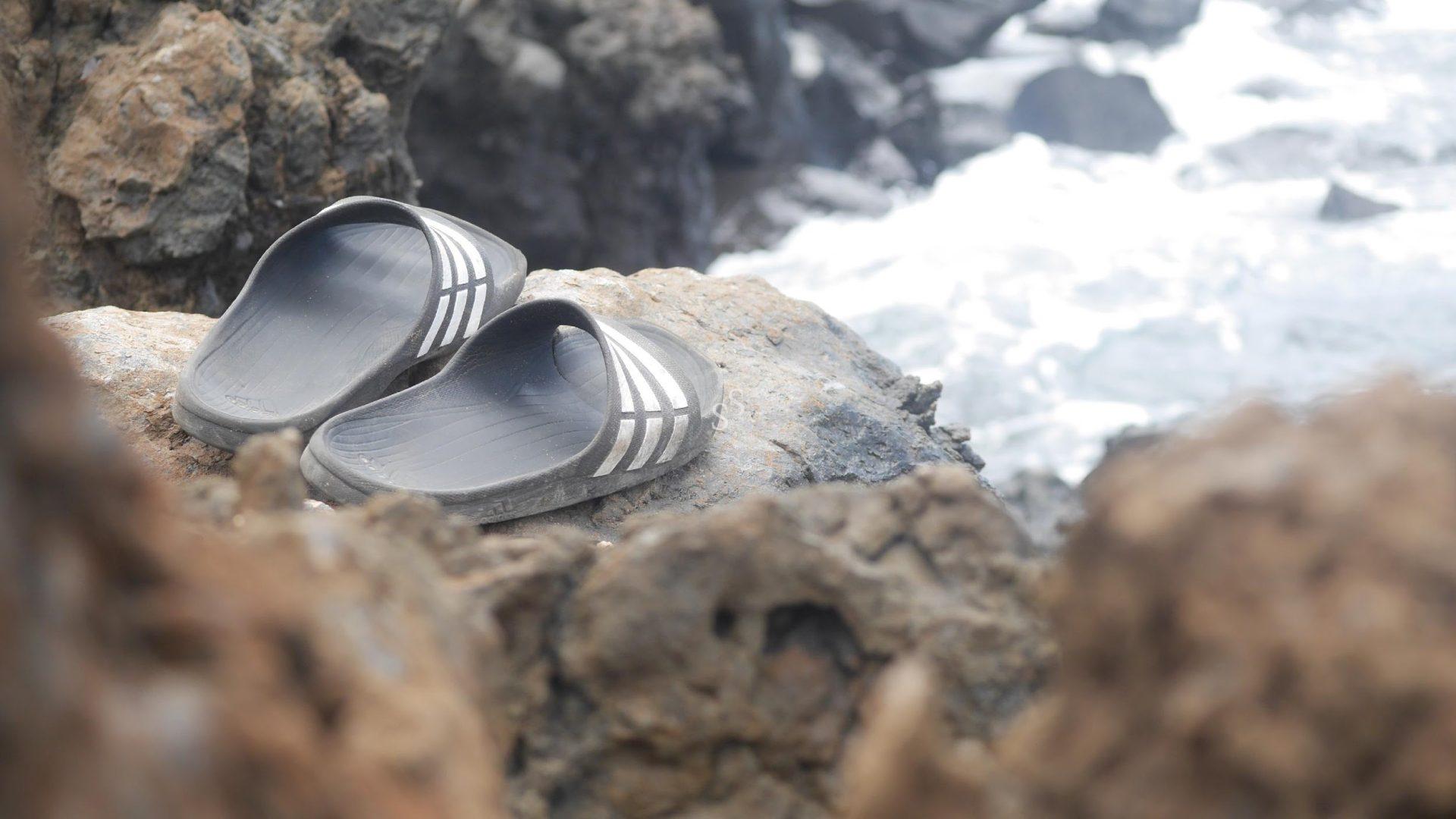 A pair of black slide sandals with white stripes rests on a rocky surface near the ocean, with waves and rocks blurred in the background.