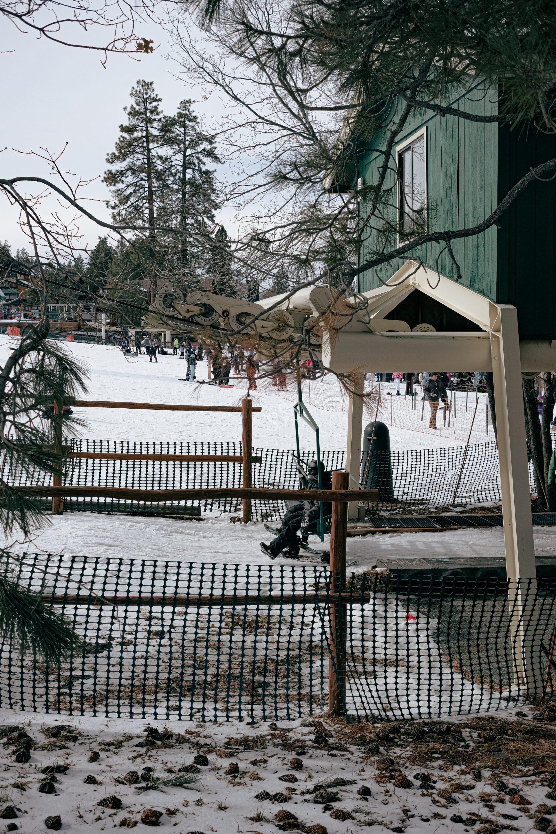 A snow-covered area with people in winter clothing, a green building with a sloped roof in the foreground, pine trees, and a wooden fence surrounding the scene.