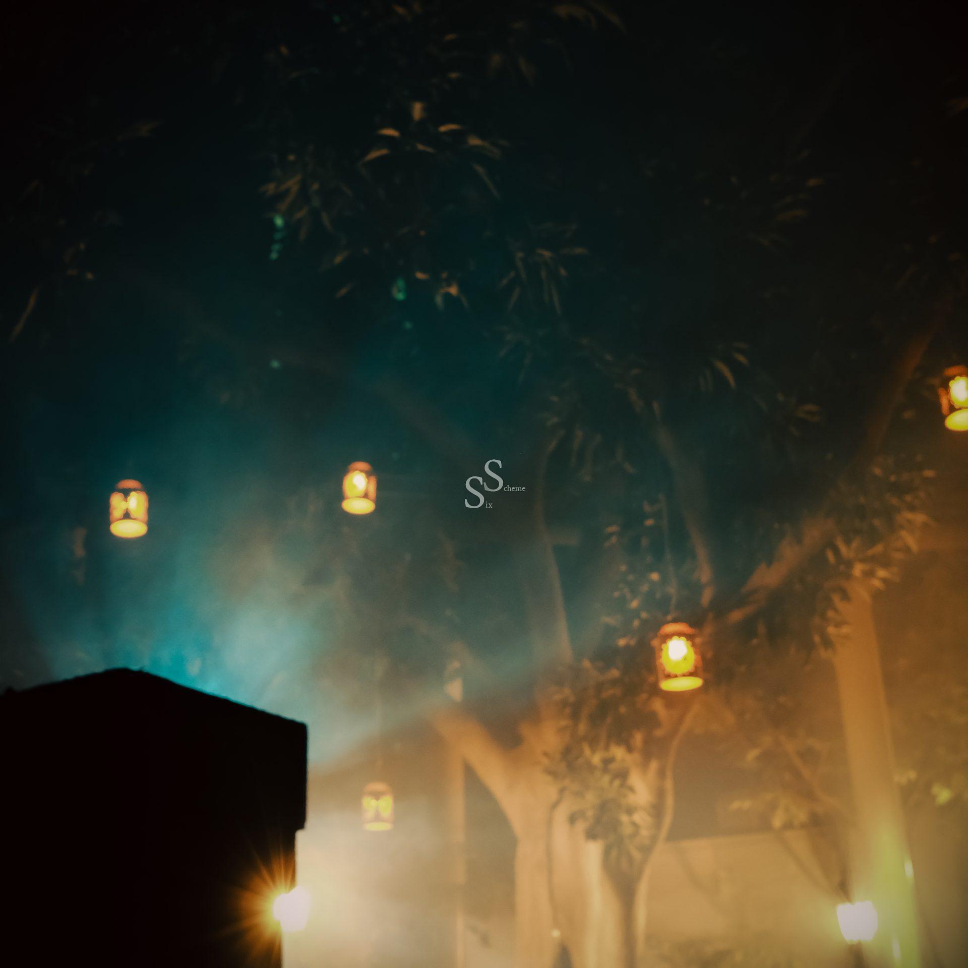 Lanterns hang from tree branches, glowing softly through a hazy, mist-filled night. The background is dreamy and atmospheric, with blue and yellow lights illuminating the scene.