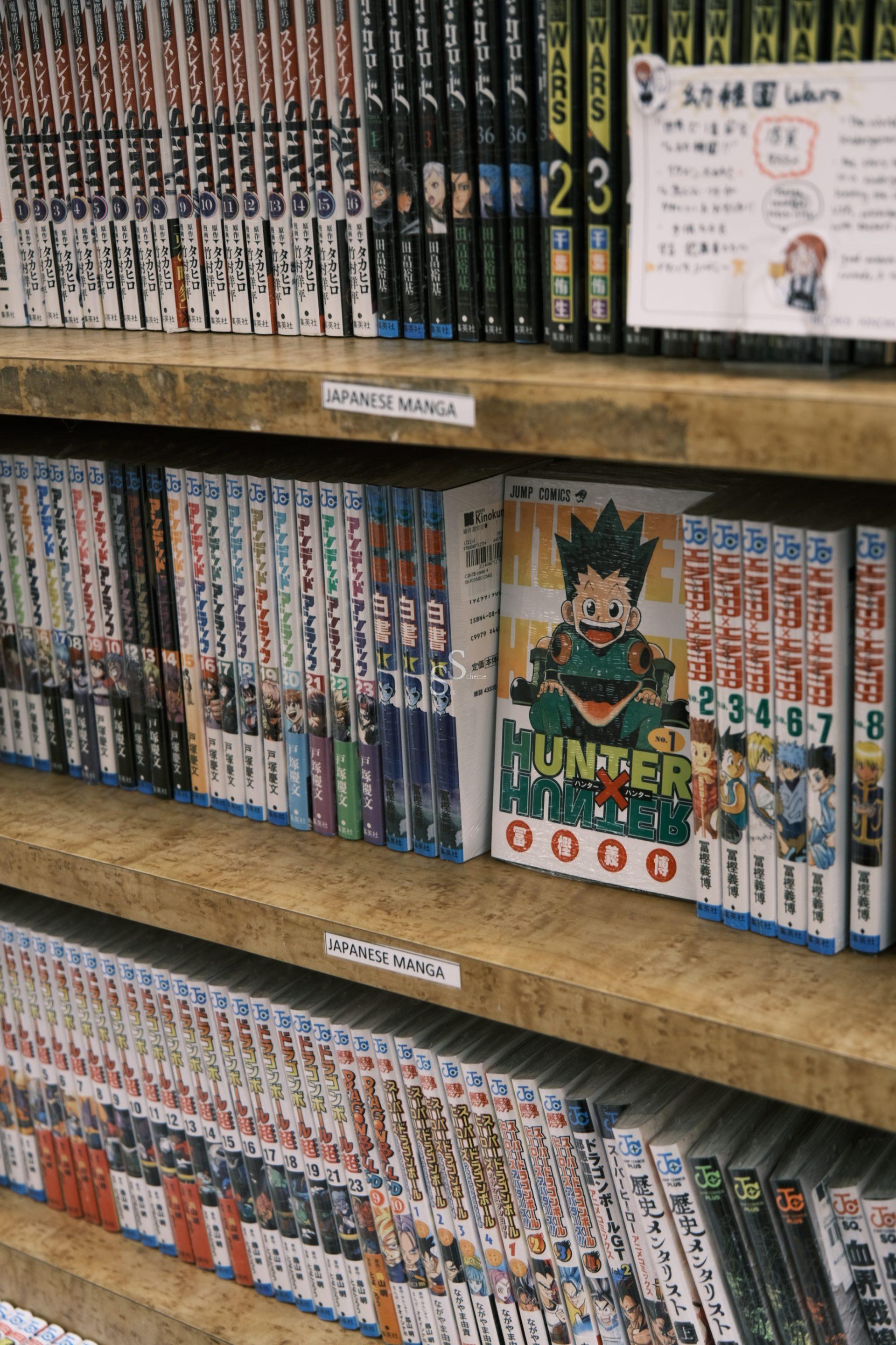 Shelves filled with Japanese manga books, neatly organized. The manga Hunter x Hunter is prominently displayed facing forward among the spines of other manga volumes. Signs label the section as Japanese Manga.