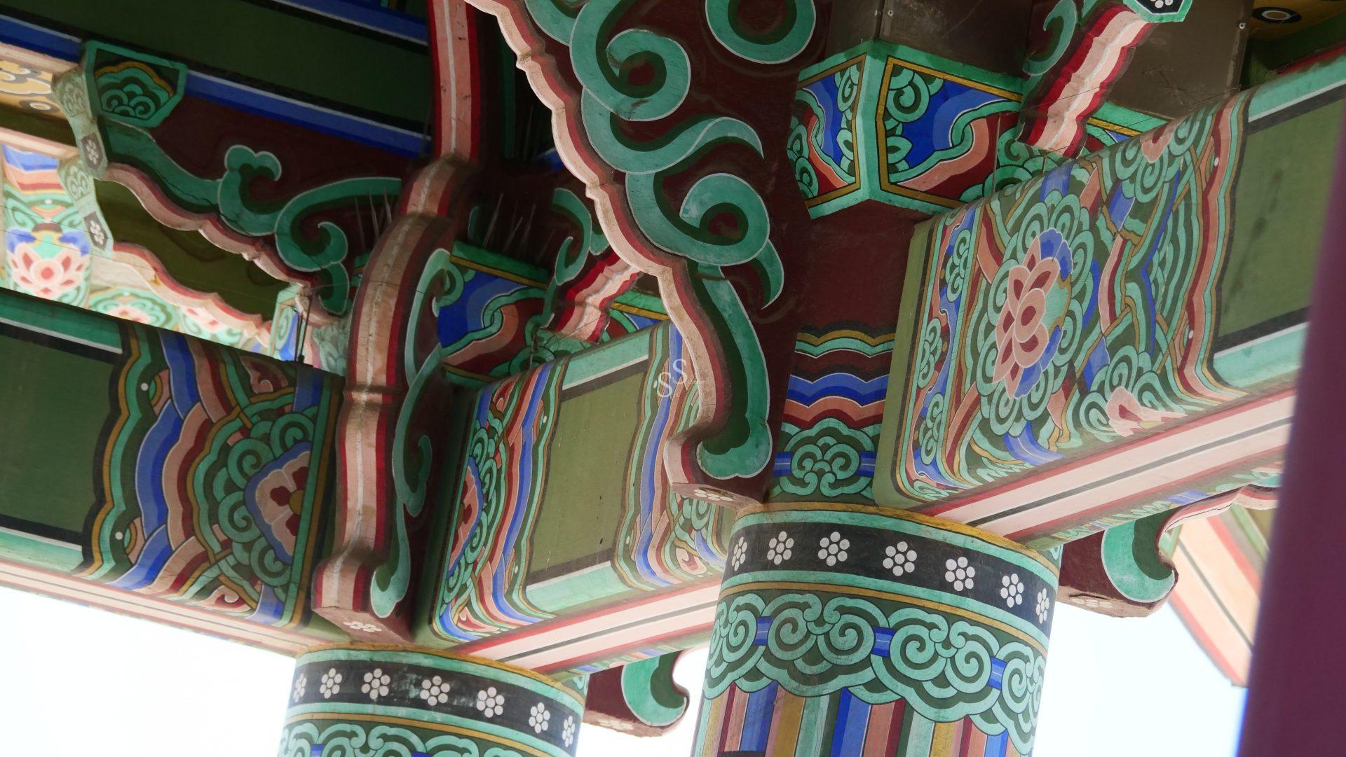 Colorful wooden beams and columns feature intricate patterns and traditional Korean designs, showcasing ornate green, red, blue, and white decorations on the underside of a historical buildings roof.