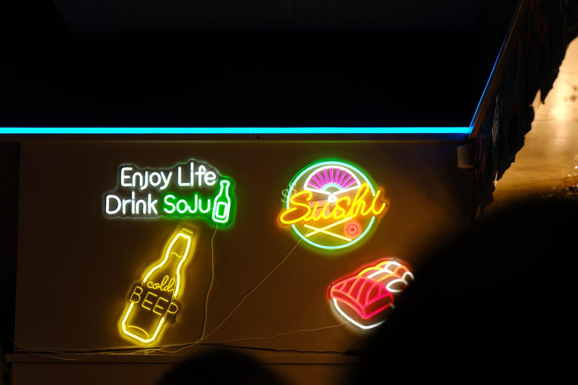 Neon signs promoting food and beverages are mounted on a wall: "Enjoy Life Drink Sake" appears with a bottle graphic, "Sushi" is shown with an icon, and a separate beer bottle sign glows nearby. The ceiling and a dark lower-right shadow are visible in the surrounding space.