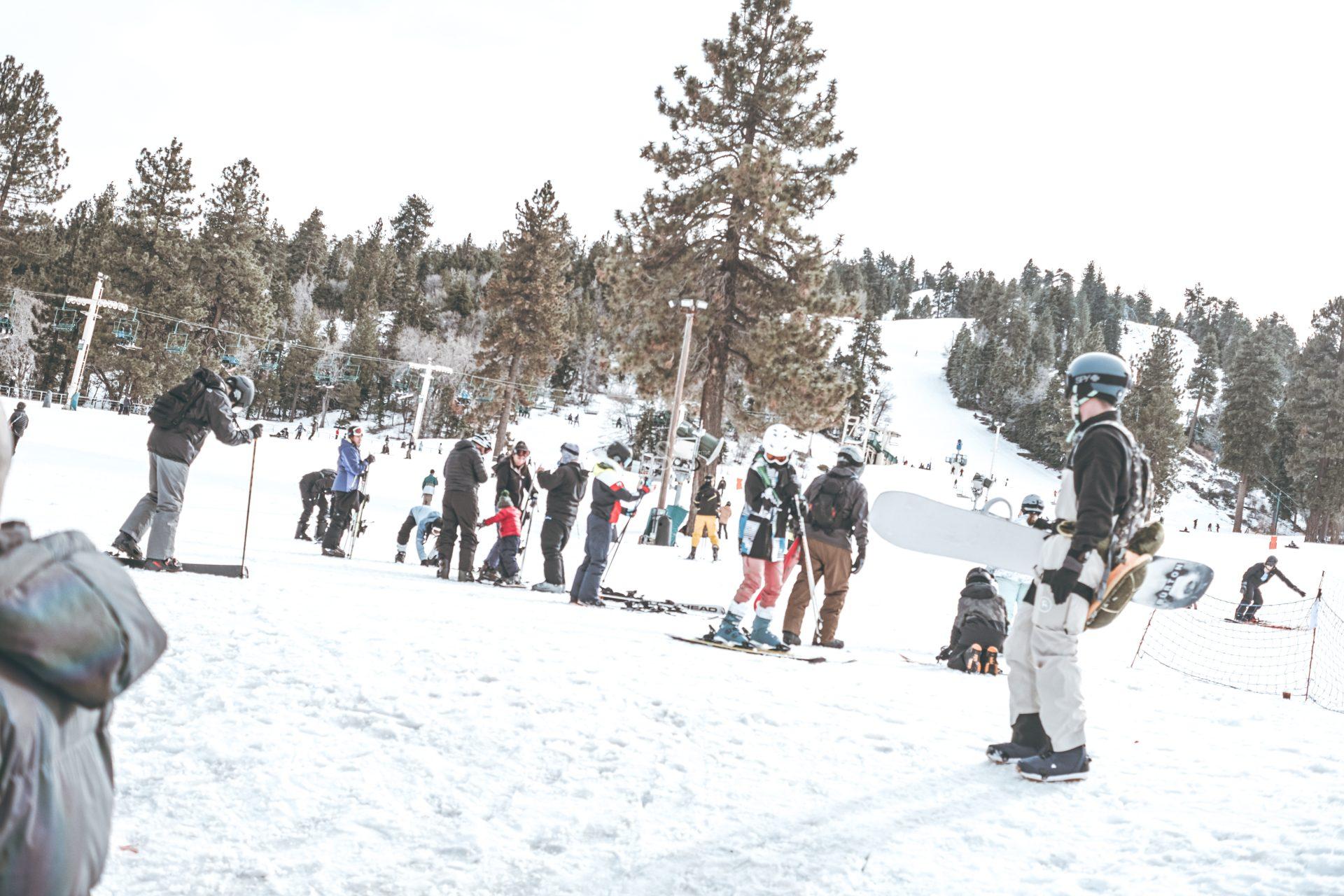 People in winter gear gather at a snowy ski resort, some holding skis or snowboards. Pine trees and ski slopes are visible in the background under an overcast sky.