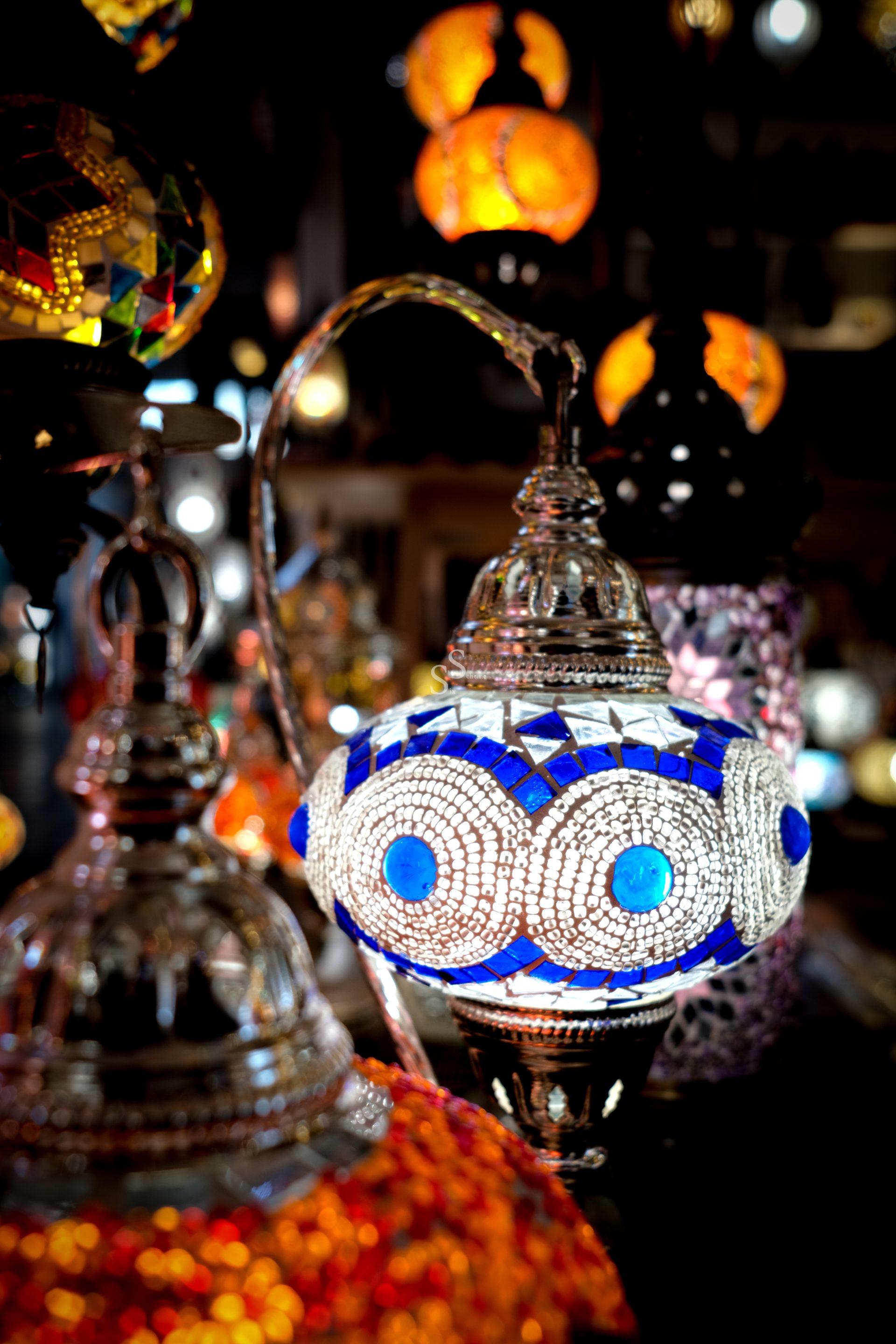 A close-up of a decorative mosaic lamp with blue and white glass pieces and intricate patterns, surrounded by other colorful, glowing lamps in the background.