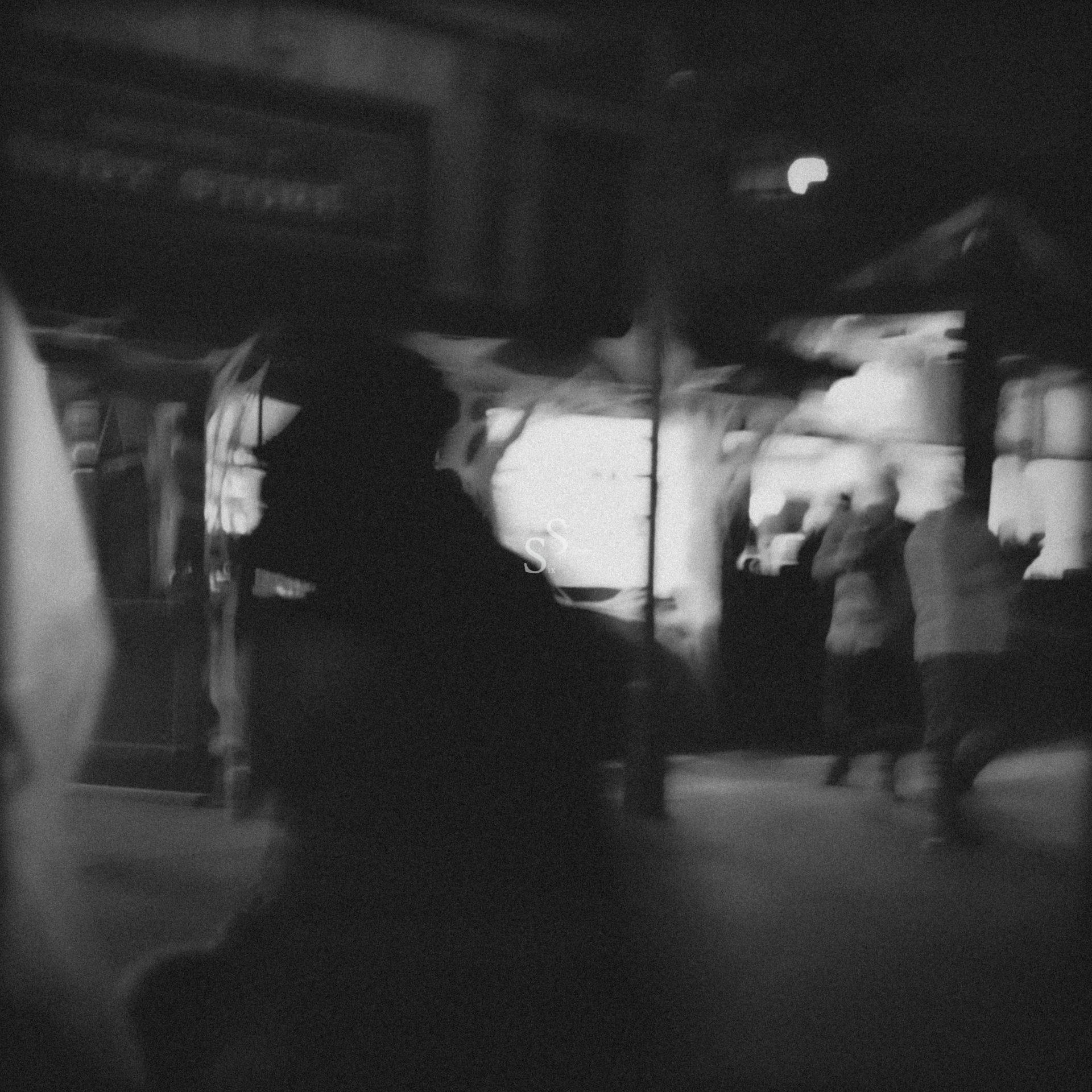 A blurry, black-and-white photo of a dimly lit street scene at night, with silhouettes of people walking and bright lights from storefronts in the background, creating an atmospheric, mysterious mood.